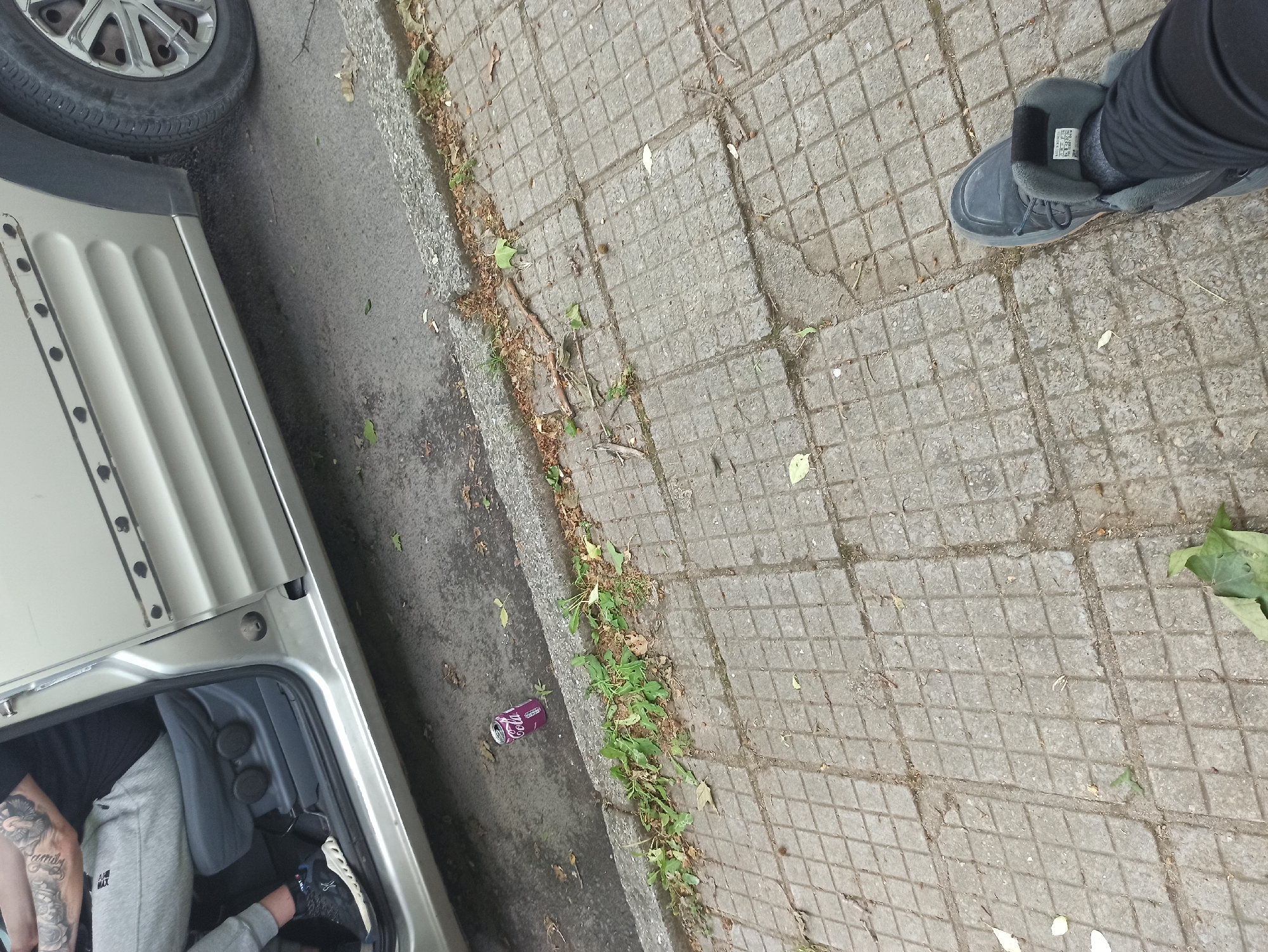 The scene shows a partial view of a city street in Unknown, Unknown. A silver vehicle is parked on the left side of the frame, with its open trunk revealing a person wearing gray sweatpants and black shoes with white soles inside. Visible tattoos are present on the person's arm. Next to the vehicle, on the street curb, there is a discarded purple Coca-Cola can amidst some weeds and debris. The sidewalk consists of gray concrete square pavers, with some leaves and dirt scattered on its surface. On the right side of the frame, a person wearing black pants and gray shoes is visible.
