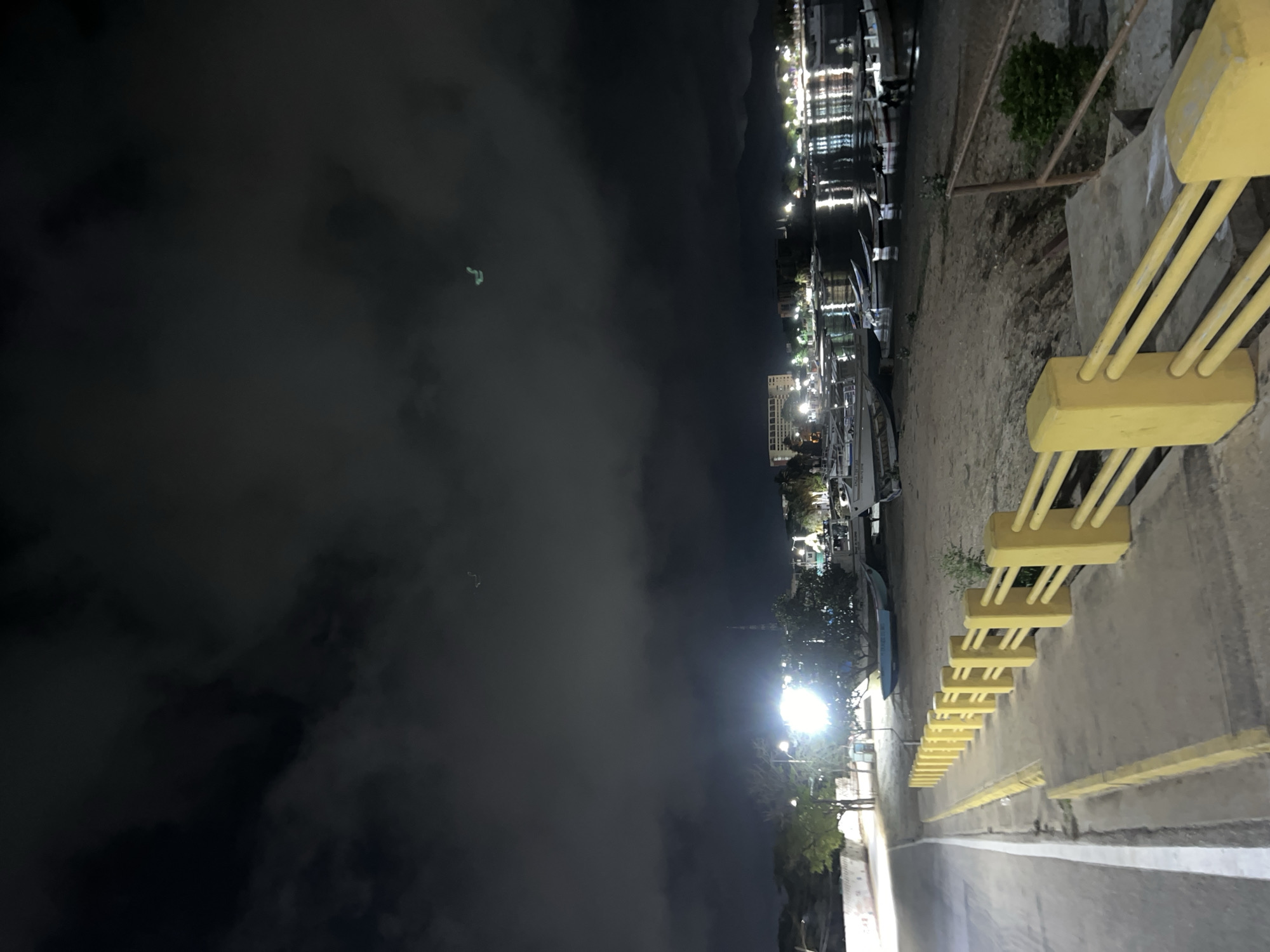 A nighttime, eye-level, wide shot captures the waterfront of Juan Griego, Venezuela. The scene is dominated by a dark, cloudy sky with scattered lights visible in the distance, reflecting on the water. In the foreground, a yellow-railed concrete barrier runs along a sandy embankment. Beyond the barrier, a body of water is dotted with numerous small boats, some of which are beached on the sand. A street runs along the left side of the image, illuminated by a bright light source in the middle ground. Buildings with lights are visible in the distance, along with distant mountains. The overall atmosphere is dimly lit and somewhat ominous due to the dark sky and limited visibility.
