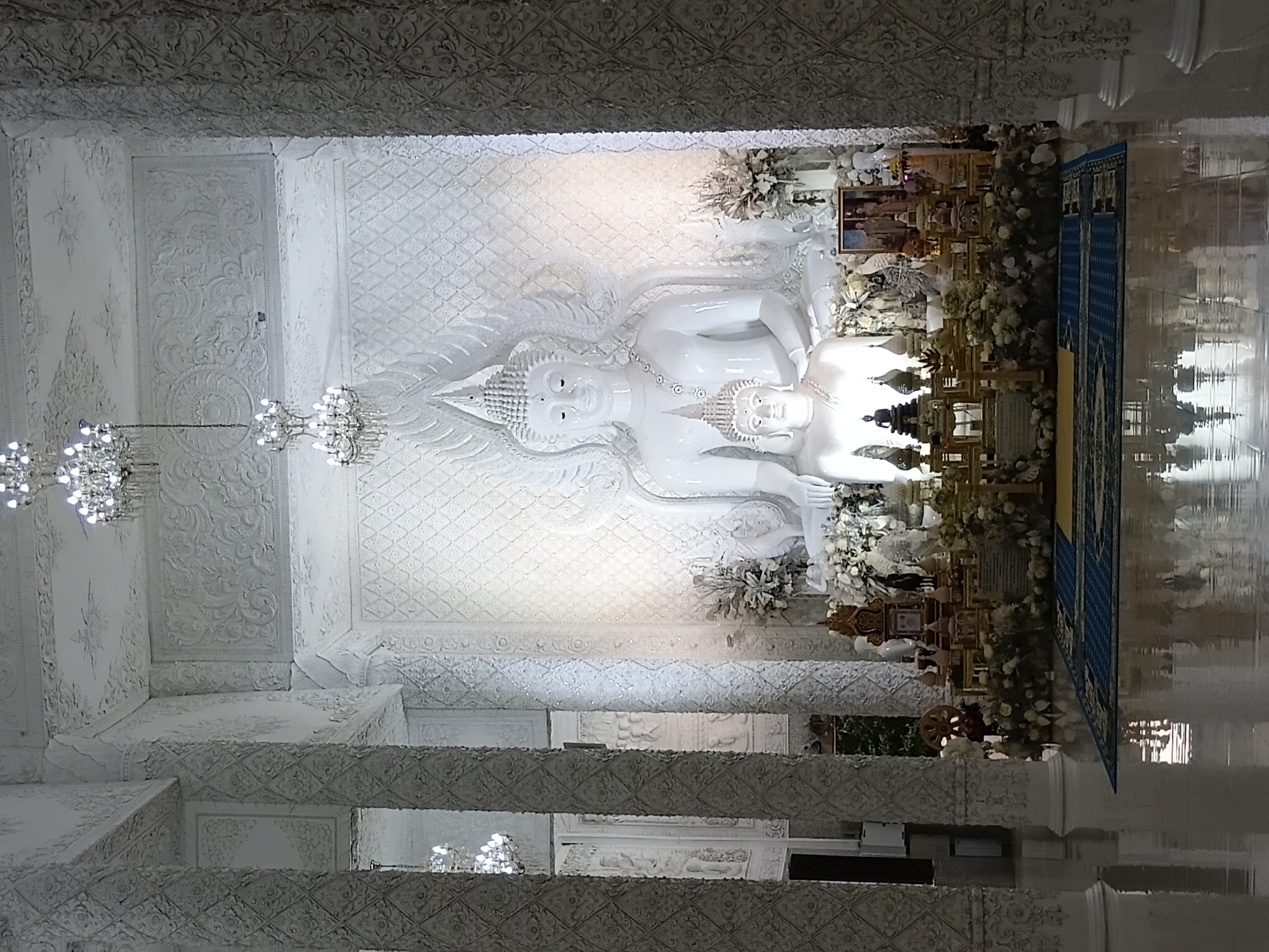 This is a full-shot, eye-level, indoor photograph of a richly decorated white Buddhist temple in Ban Lao Phatthana, Thailand. The primary subject is a large, white, seated Buddha statue with another smaller Buddha statue in its lap. Both statues are adorned with crowns and necklaces. The statues are positioned in front of a white, ornate wall with intricate carvings and a larger, raised carving of a Buddha head at the apex.
Surrounding the statues is a raised altar adorned with numerous small, tiered structures, some topped with spires, and arrangements of white flowers. A framed photograph depicting two individuals is also visible on the altar. In front of the altar, a blue and yellow patterned mat is laid out on a highly reflective white floor.
The scene is flanked by large, white, ornately carved pillars that extend upwards towards a ceiling with elaborate plasterwork and decorative elements. Two crystal chandeliers hang from the ceiling, illuminating the space with bright, cool light. The overall impression is one of opulence, serenity, and spiritual significance. The reflective floor creates mirror-like images of the altar, statues, and pillars, adding to the ethereal quality of the scene.