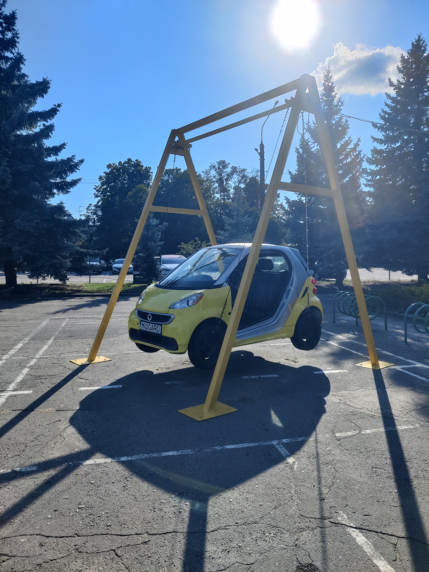 A yellow Smart car is suspended mid-air from a yellow metal swing set structure in an outdoor parking lot. The car is positioned with its front facing the viewer, and its doors are open. The swing set has two triangular supports on each side, anchored to the asphalt ground with metal plates. The parking lot has white painted lines, and in the background, there are trees, other parked vehicles, and bicycle racks. The sun is shining brightly from the top right of the frame, casting long shadows of the swing set and the car onto the ground. The sky is clear and blue. The license plate of the car reads "MIDGARD". The scene appears to be located in a public space, possibly a park or a street in Kyiv, Ukraine, given the context of the user's request.