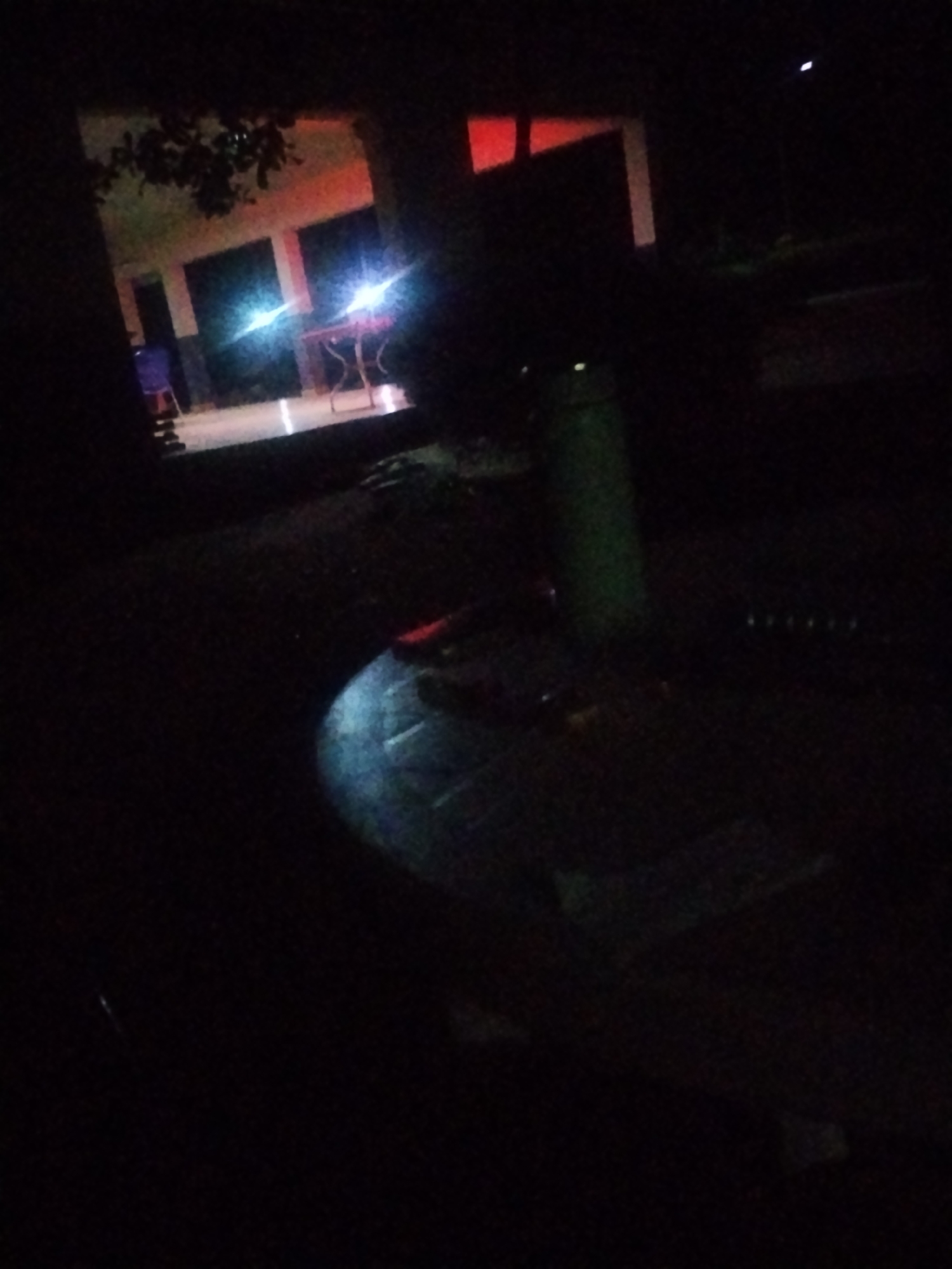 The image depicts an outdoor scene at night in Mangu Town, Nigeria. The setting appears to be a courtyard or paved area adjacent to a building. In the background, there's a building with illuminated windows showing a reddish-orange glow, possibly from interior lighting or decorative lights. Two bright white lights, potentially from car headlights or powerful lamps, are prominently visible, casting reflections and illuminating the scene.
In the foreground, a large, dark, circular object with a textured surface is visible, taking up a significant portion of the frame. Its exact nature is unclear due to the low light conditions and the image's resolution, but it could be a large pot, a decorative planter, or a piece of outdoor furniture. To the left of this object, a slender green cylindrical object, possibly a post or a narrow container, stands upright.
The overall mood is dark and somewhat mysterious, illuminated primarily by artificial lights. The time of day is clearly night, and there are no indications of the weather. The scene is likely a public or semi-public space, given the building and the paved area. No people are clearly discernible, and no text is visible in the image. The presence of the building suggests it might be a school, a community center, or a residential complex. The bright lights could indicate activity, or simply be ambient lighting for the area.