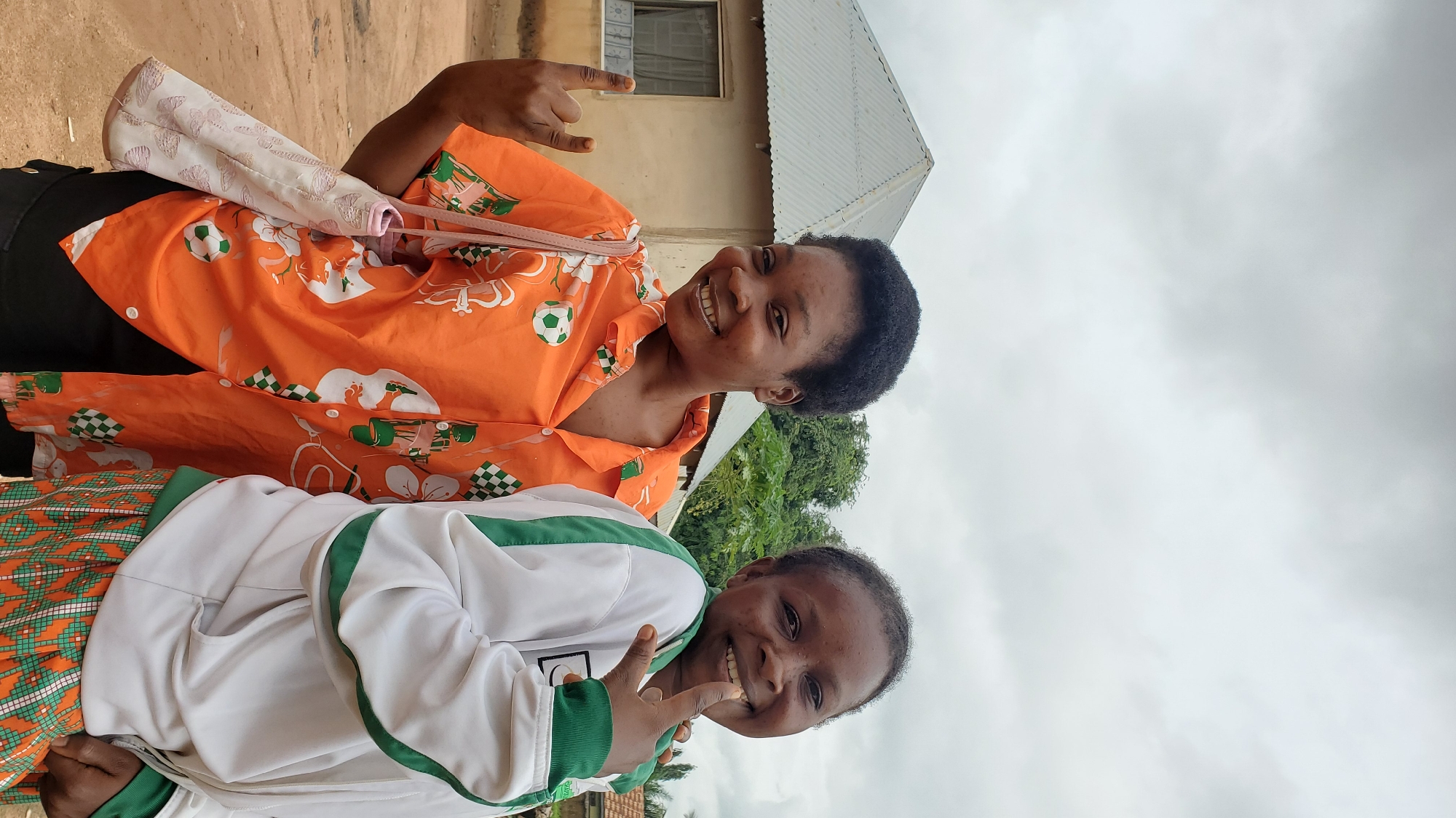 Two young women are standing in front of a modest house in Jalingo, Nigeria. The woman on the left is wearing an orange shirt with a pattern of soccer balls and flowers, and she is holding a bag on her shoulder. She is making a "rock on" hand gesture. The woman on the right is wearing a white jacket with green trim, and she is pointing her finger up at the camera. The house behind them is simple, with a corrugated metal roof and a window. The sky is overcast.