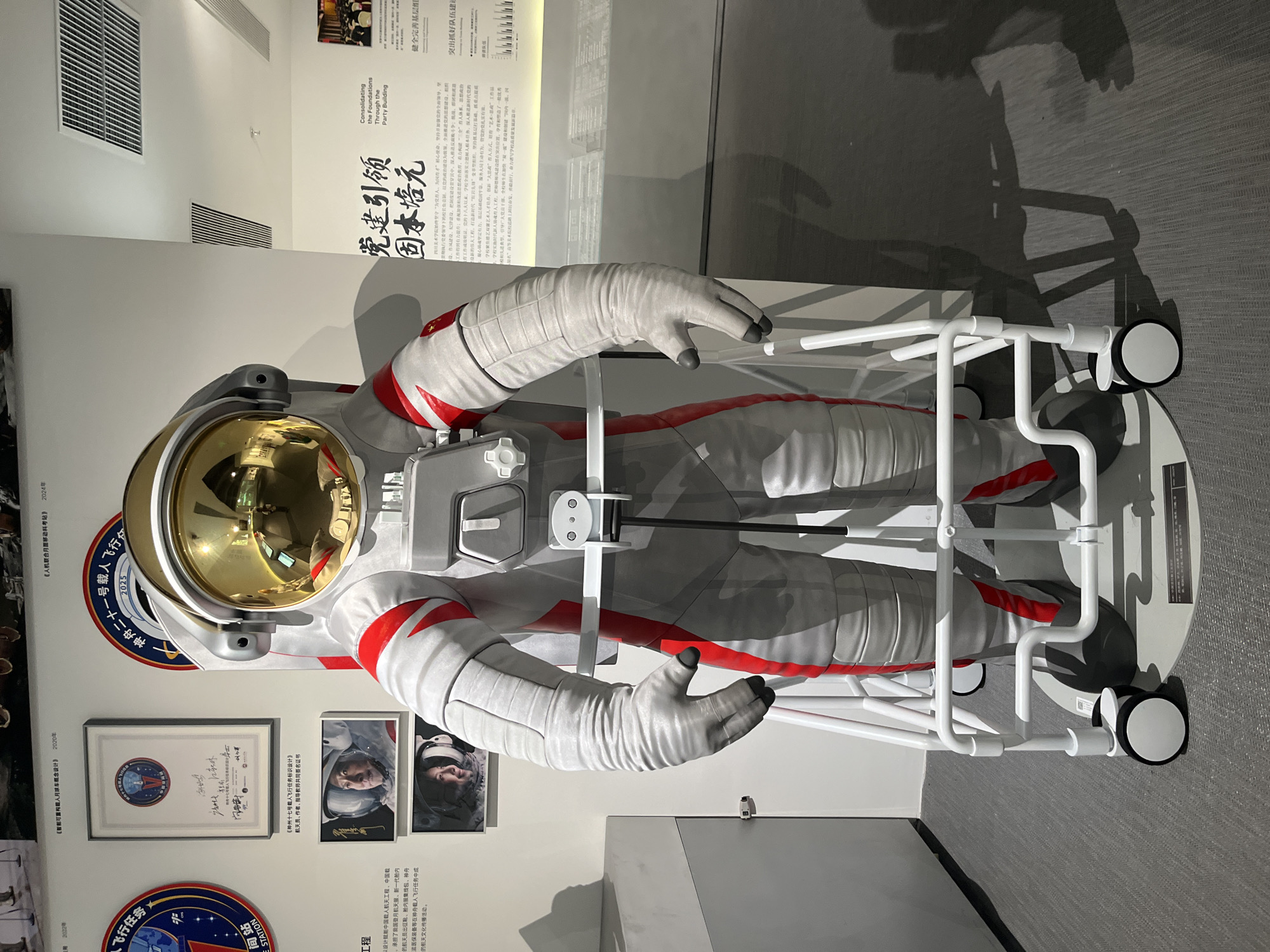 An astronaut suit is displayed on a white wheeled stand inside an exhibition hall. The suit is predominantly white with red accents and a golden helmet. The exhibition hall appears to be a museum, with various displays and informational panels on the walls. One panel visible in the image is written in Chinese. The floor is carpeted in a gray speckled pattern. The location is the Chongqing High-tech Zone, China.