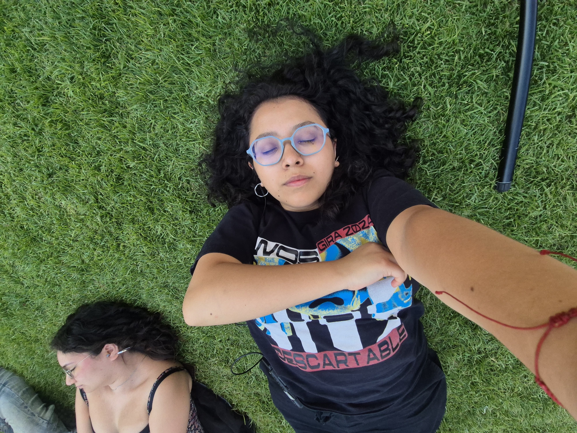 Two women are lying on a green grassy lawn. The woman in the foreground is lying on her back, facing the camera. She has curly dark hair, wears light blue-rimmed glasses, and has a black t-shirt with a graphic design. Her arms are folded across her chest, and she appears to be resting with her eyes closed. Her right arm extends towards the camera, adorned with a red string bracelet. To the left of the main subject, partially visible, is another woman also lying down. This woman has dark curly hair and wears a dark top with floral patterns and glasses. A black cylindrical object is visible near the first woman. The grass appears to be manicured. It's difficult to ascertain the exact location in Puebla, Mexico, without more specific details.