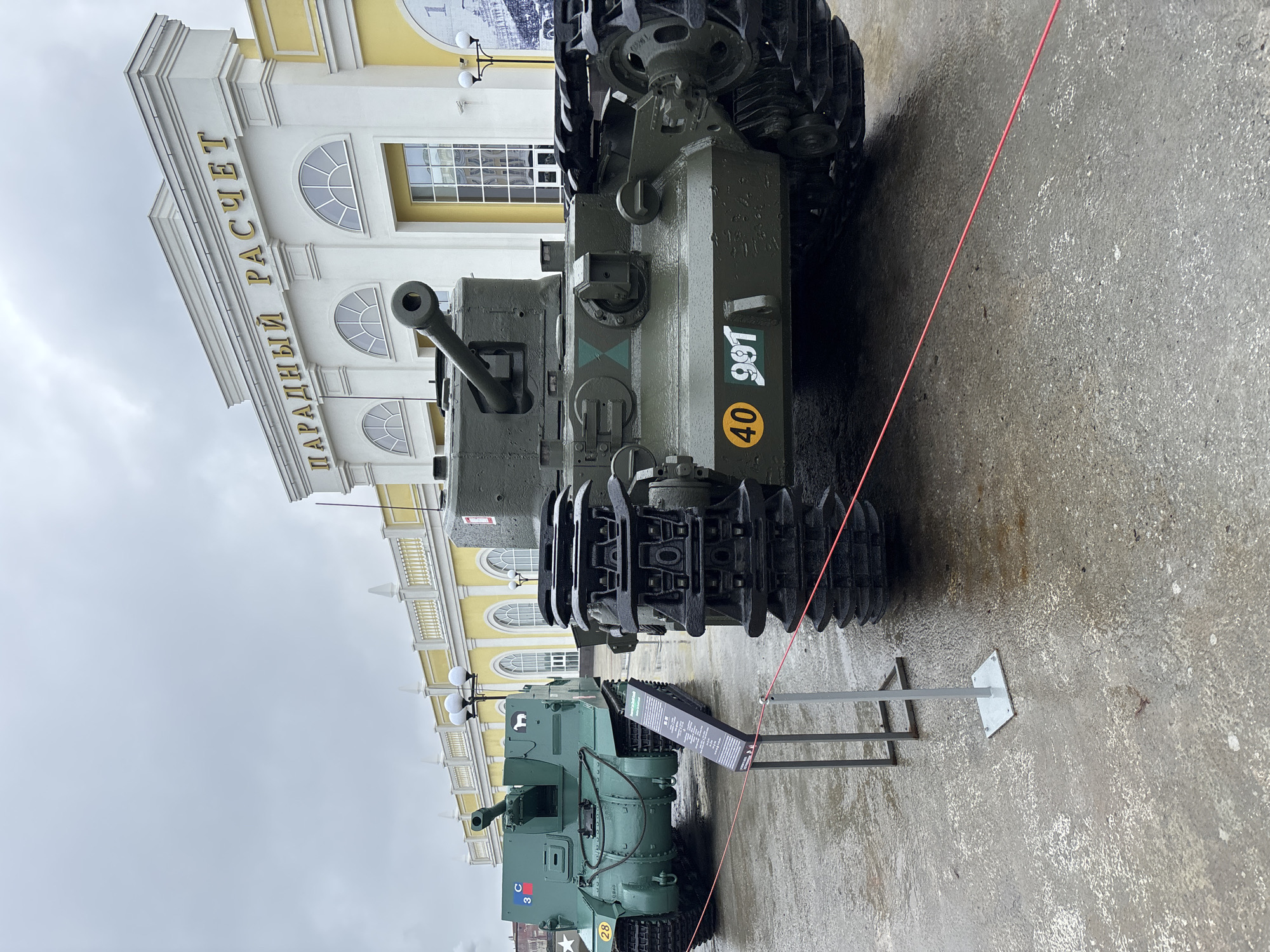 In Verkhnyaya Pyshma, Russia, two military vehicles are on display in front of a yellow building with the words "ПАРАДНЫЙ РАСЧЕТ" (Parade Calculation) in gold lettering. The vehicle in the foreground is a dark green tank with black tracks. It has a yellow circle with the number 40 and white numbers 991 on its side. The vehicle behind it is also green and appears to be an artillery piece or a self-propelled gun, with a designation of "C3" and a white star emblem on its side. The ground is wet and appears to be asphalt or concrete, suggesting recent rain. A red rope is stretched across the ground in front of the tanks, serving as a barrier. There is a small informational plaque in front of the second tank. The sky is overcast.