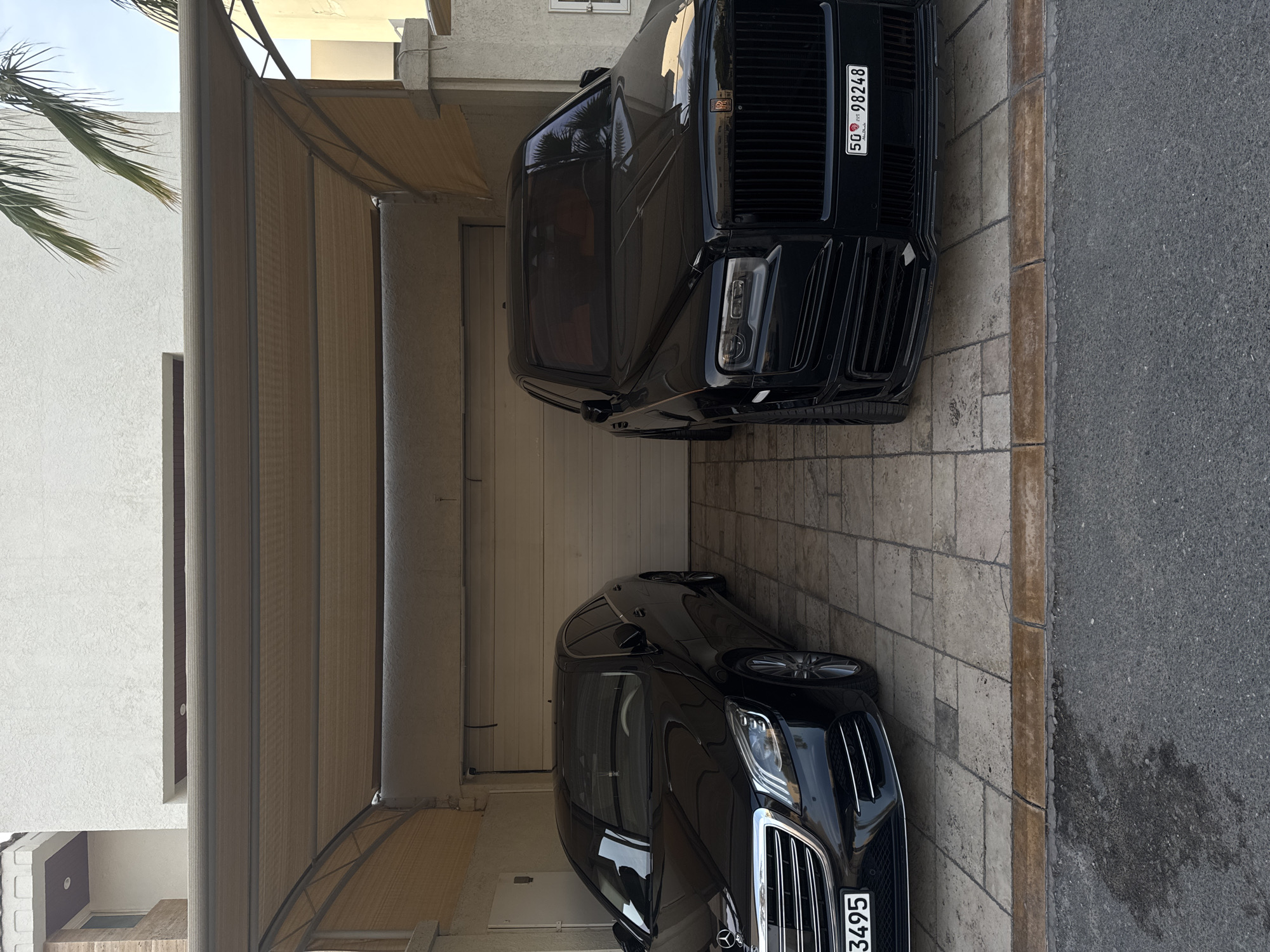 The image shows two luxury cars parked in a garage or carport. The top vehicle is a black Rolls Royce, identifiable by its distinctive front grille and the license plate "50 98248". The bottom vehicle is a black Mercedes-Benz, discernible by its grille and the license plate "...3495". The vehicles are parked on a beige-colored stone-tiled surface. The garage is covered with a retractable beige awning, supported by vertical posts. A residential building is visible on the left side of the frame. Based on the license plates, the location can be inferred to be Dubai, United Arab Emirates.