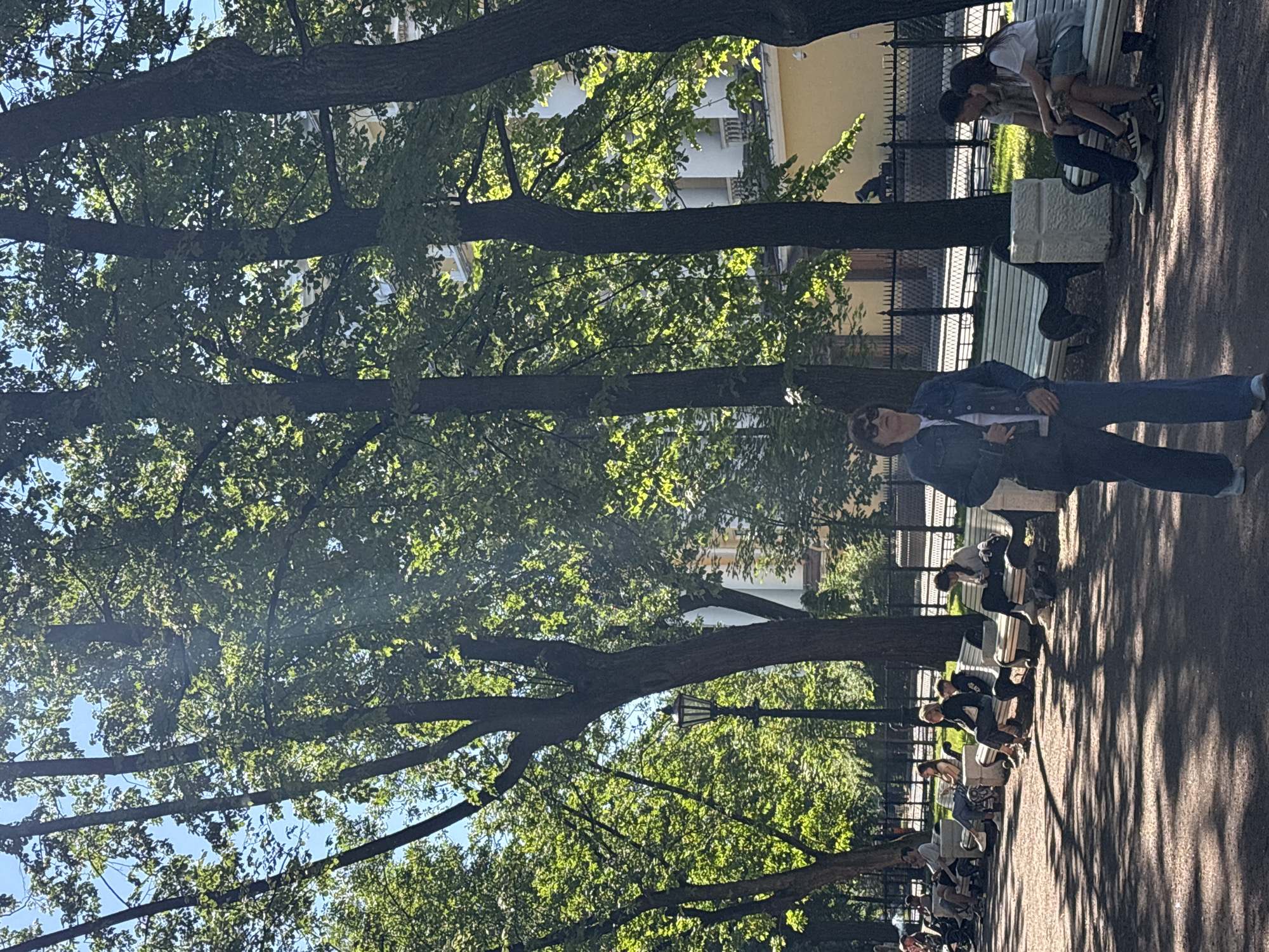 The image shows a sunny day in a park in Saint Petersburg, Russia. In the foreground, a woman walks towards the viewer. She is wearing a denim jacket and blue pants. Behind her, several people are sitting on benches. To the right, a couple is sitting close together on a bench, and the woman is looking at her phone. To the left, more people are sitting on benches, some reading or looking at their phones. The park features tall trees with green foliage, casting shadows on the paved walkway and benches. A street lamp is visible between the trees. In the background, a building with classical architecture, featuring white columns and yellow walls, can be seen behind a black metal fence. The overall scene depicts a leisurely day in a public park.
