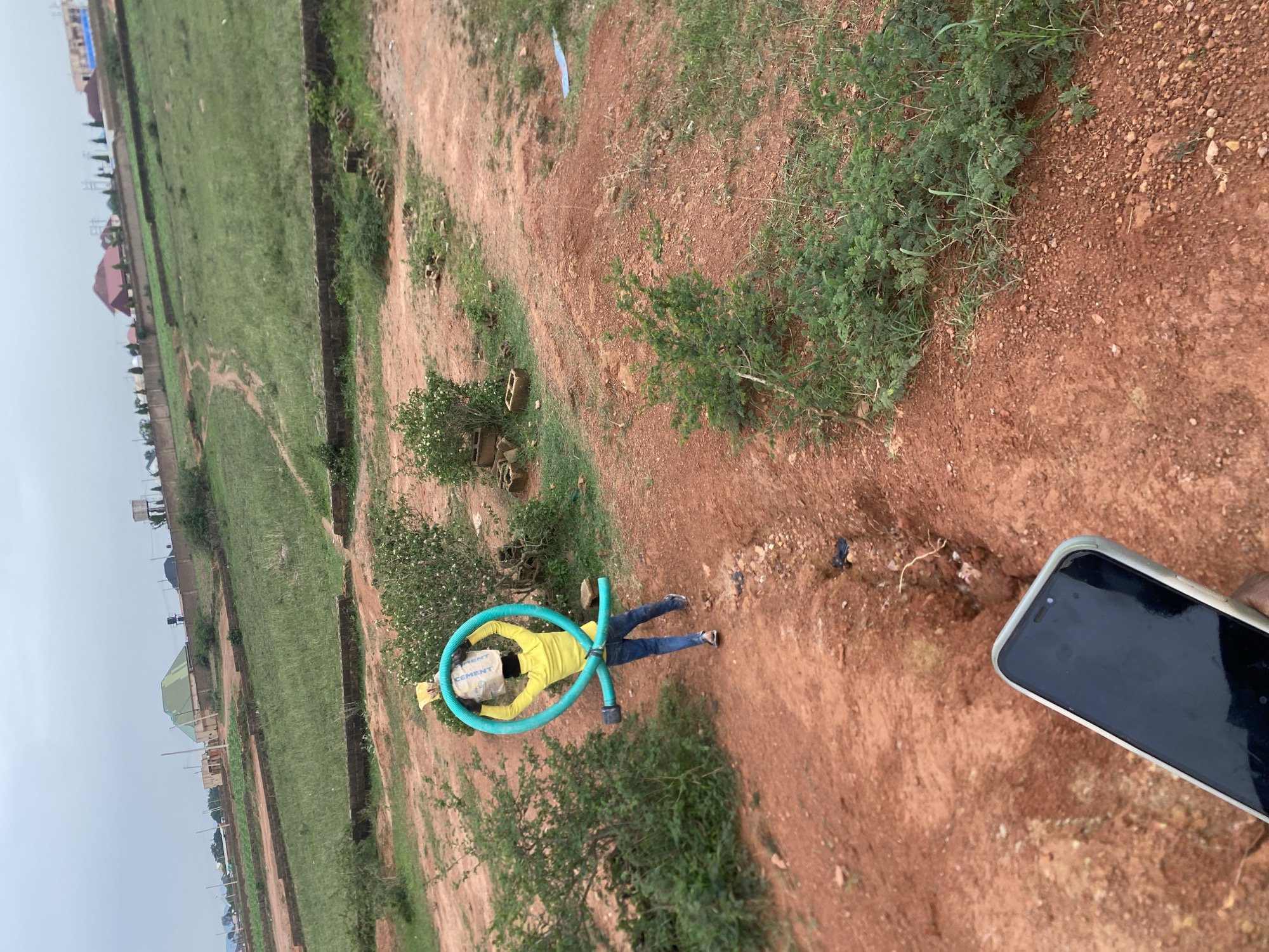 A person wearing a yellow shirt and blue jeans is walking away from the camera on a dirt path. They are carrying a large bundle wrapped in a yellow plastic bag on their head, and a green coiled pipe is around their head, resting on the bundle. The person is in a hilly, undeveloped area with sparse vegetation and scattered bricks. In the background, there are some buildings, a water tower, and a walled compound. This scene is consistent with a construction or development site in the rapidly expanding outskirts of Jos, Nigeria. A hand holding a dark smartphone is visible in the foreground, obscuring part of the scene.