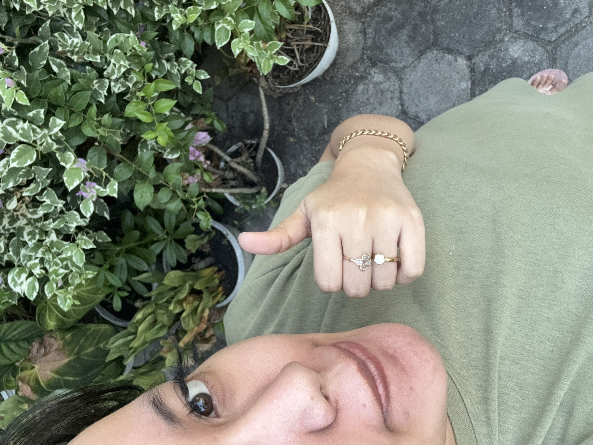 In Gadingrejo, Indonesia, a close-up, high-angle, outdoor shot shows a person wearing a green t-shirt. Their left eye, with long eyelashes and an eyebrow, is visible in the upper left of the frame. The person's mouth is partially obscured. Their left hand is raised, giving a thumbs-up gesture, and adorned with two rings: one appears to be a clover design with small stones, and the other is a simple band with a single stone. A gold chain bracelet is worn on their wrist. The background consists of potted plants, some with green and white variegated leaves, and others with solid green foliage. The ground is paved with dark gray, irregularly shaped stones.
