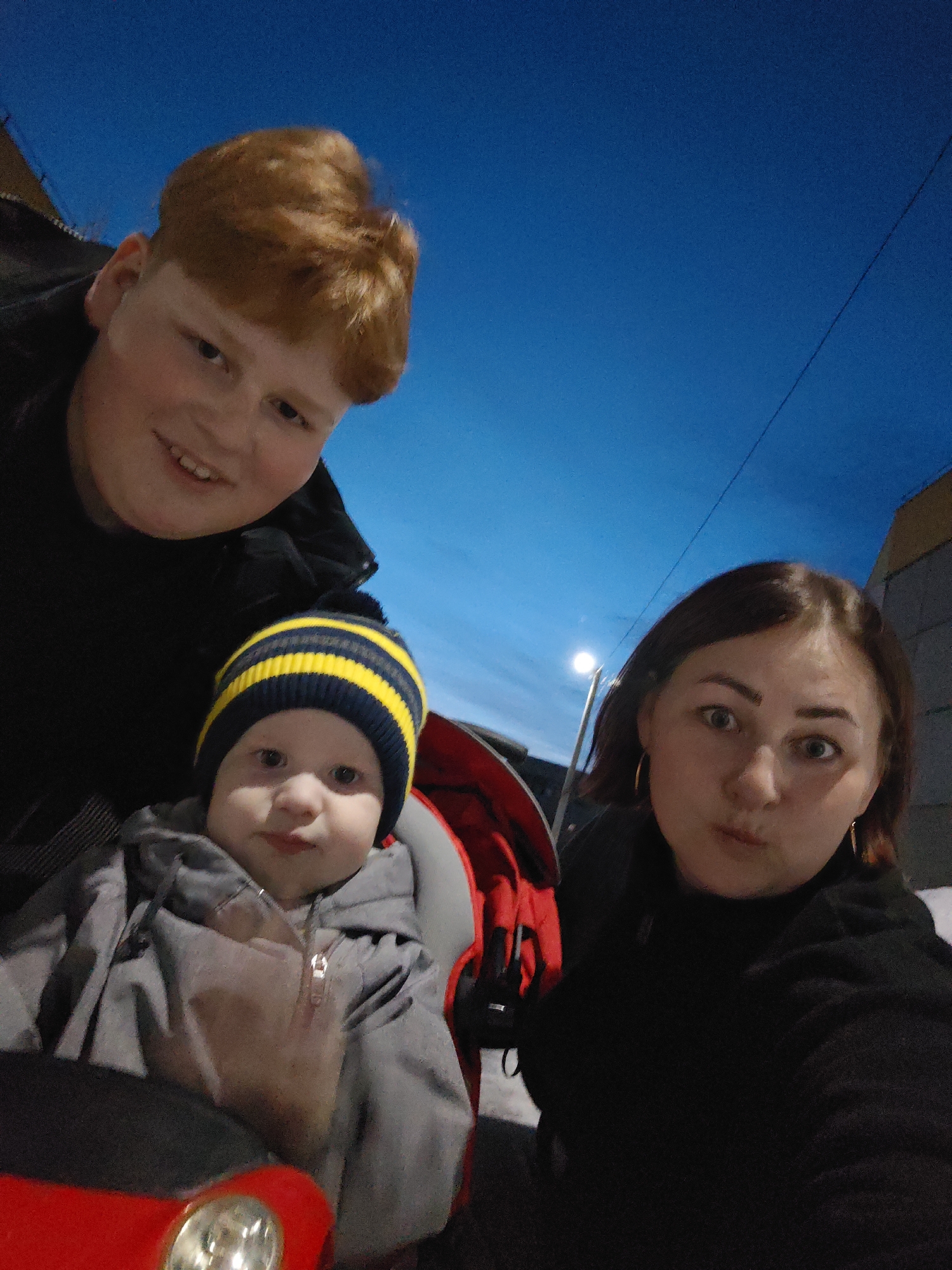 An upward-angled photograph captured in Марковский, Russia, depicts three individuals outdoors at dusk or night. In the upper left quadrant, a male with reddish-blonde hair and a dark jacket smiles directly at the camera. Centered below him is an infant or young child, wearing a grey hooded jacket and a navy and yellow striped beanie, seated in a red and grey child transport device, also looking directly forward. To the right, an adult female with dark brown hair and gold hoop earrings looks intently at the camera with widened eyes and pursed lips. The background features a deep blue sky. To the far right, a light-colored building is partially visible. A street lamp, illuminated, stands upright in the mid-ground behind the female, with a dark wire crossing the sky above her. Snow is faintly visible on the ground in the mid-ground. The lower left foreground shows the red front portion of the child's transport device, including a metallic element.