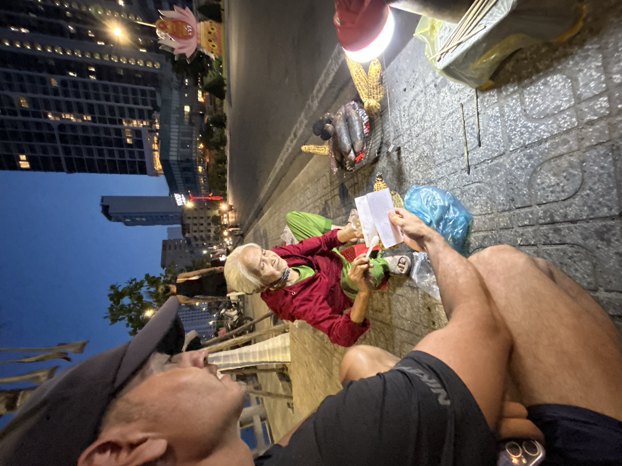 An eye-level shot with a slightly wide-angle lens captures a street scene in Bắc Nha Trang, Vietnam. An elderly woman sits on the sidewalk on a low makeshift stool. She wears a red jacket over a green shirt and light green pants, and sandals. She appears to be handing something to the person taking the photograph, who is seated on the sidewalk with legs crossed. The person is wearing a black t-shirt and a grey baseball cap. A mobile phone is visible next to the seated person. Between them is a blue plastic bag and several objects are strewn about. To the right of the elderly woman is a small charcoal grill on which several squid are roasting. Above the grill hangs a red work light. To the right of the grill are various items, including corn on the cob and what appear to be chopsticks in a container. Behind the sidewalk is a busy street with several tall buildings in the background. The sky is dusky with hints of blue and orange. A large lotus blossom decoration adorns one of the buildings. The scene appears to be taken at dusk or early evening.