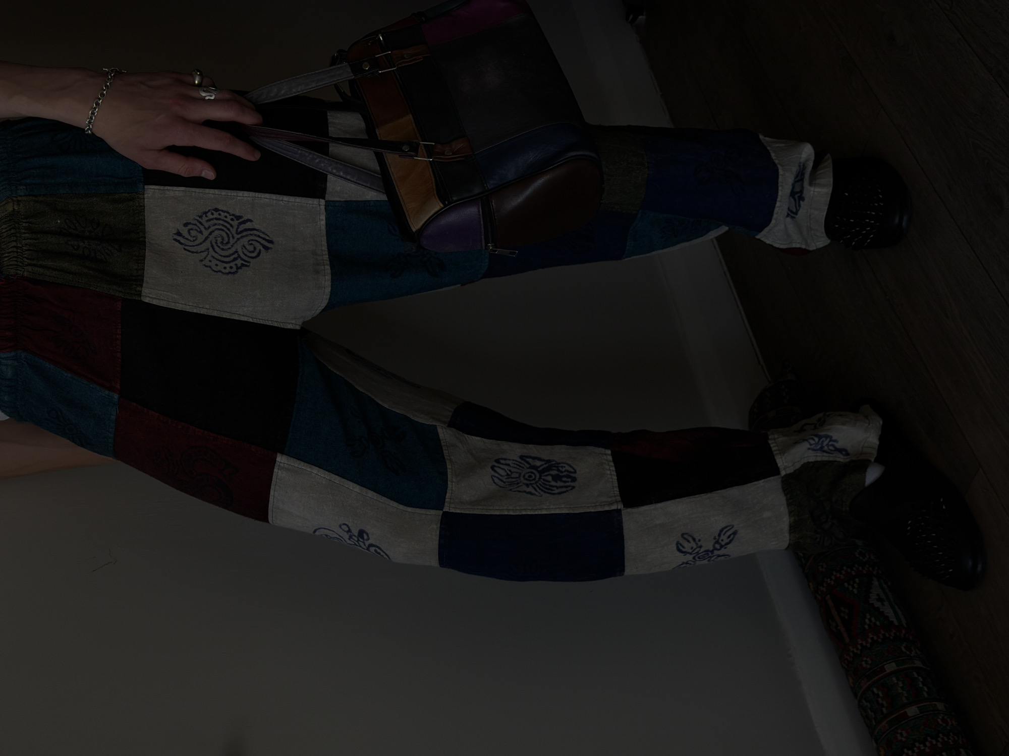 The image depicts a person wearing patchwork pants and black shoes. The pants are made of various square patches with different colors and patterns, some featuring printed designs. A multicolored leather handbag rests on the person's left leg. The person has a bracelet and a ring on their left hand. The setting is indoors, with a portion of a light-colored wall visible. A section of wooden flooring can be seen near the person's right foot. The overall lighting is dim. There is no indication of the location being in the city of London, United Kingdom.