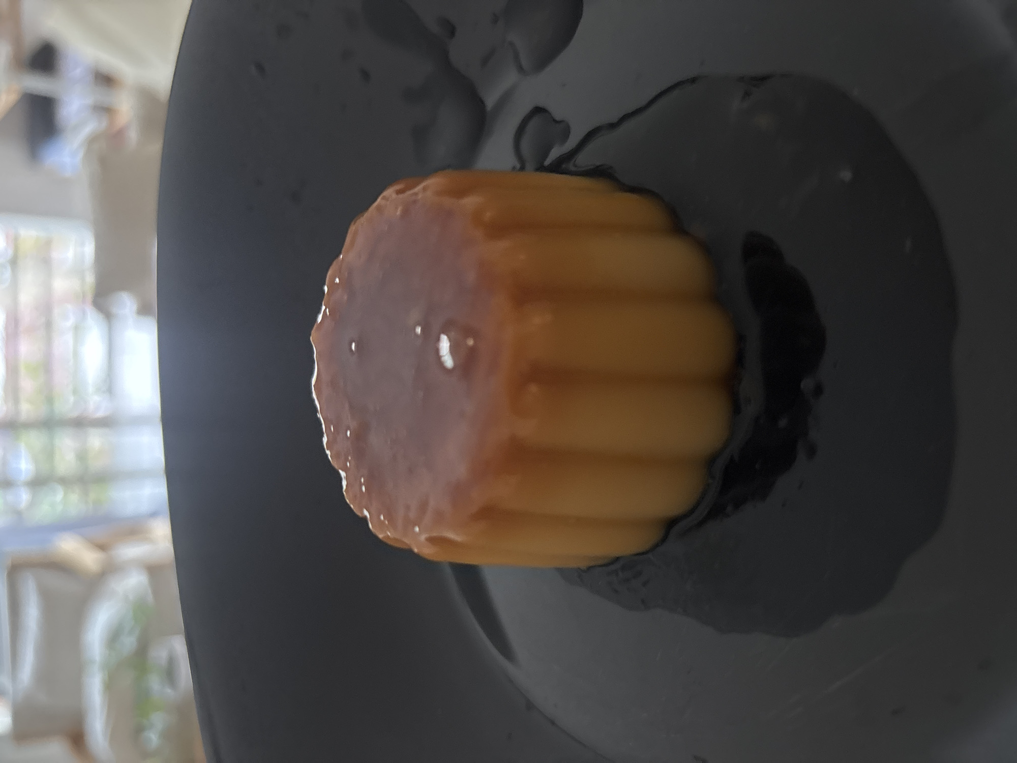 A creme caramel dessert sits on a gray plate. The dessert has a caramelized top and ridged sides. A clear syrup surrounds the dessert on the plate. A blurred background is visible through a window, with light filtering through. The location is likely a domestic setting. The scene does not include the city of Buenos Aires, Argentina.