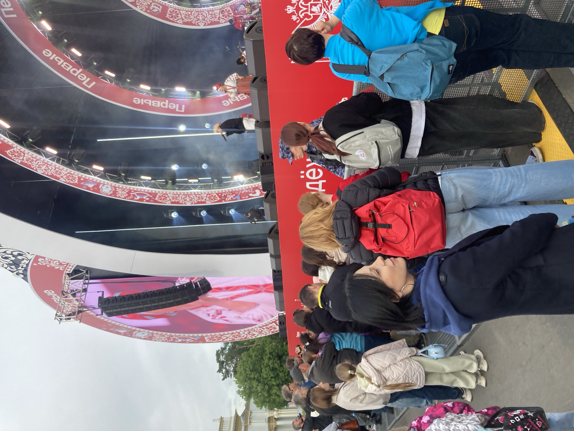 A medium, slightly high-angle shot depicts a crowd of people in Moscow, Russia, likely attending a concert or event. The crowd is primarily composed of adults, with visible variations in attire, including casual wear, backpacks, jackets, and sweaters. They are facing a stage that features large screens displaying images, with performers visible on the stage itself. Russian signage with a floral pattern adorns the stage structure. The environment includes trees and white, classical building architecture. The weather appears overcast.
