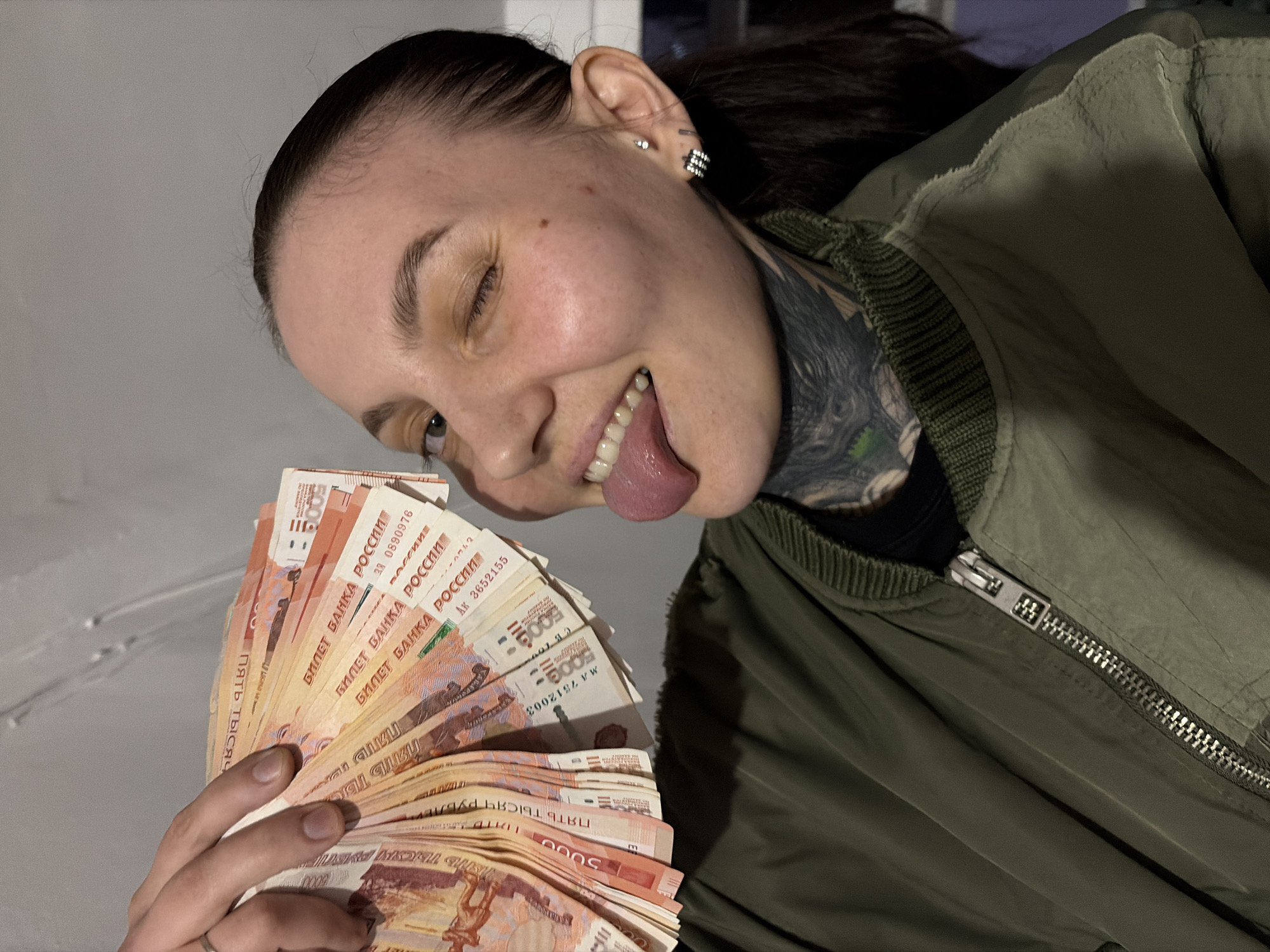The image displays a close-up, chest-up shot of a young adult female indoors in Chaikovsky, Russia. She has dark hair, fair skin with a visible mole on her right cheek, and is wearing an olive-green bomber jacket over a dark shirt. An extensive black-and-grey tattoo, featuring green accents, is visible on the left side of her neck, extending beneath the jacket's collar. Her right ear has multiple piercings, including a stud and two stacked, embellished hoop earrings. The individual is captured with her right eye winking and her tongue extended, indicating a playful or provocative expression. Her left hand, adorned with a silver-colored ring on the ring finger, holds a fan of approximately 10-15 Russian 5000 Ruble banknotes. These banknotes are reddish-orange and yellow-cream, featuring text such as "БИЛЕТ БАНКА РОССИИ" and "ПЯТЬ ТЫСЯЧ РУБЛЕЙ." Partially visible serial numbers include "3Я 0690976" and "AK 3652155." The background is a plain, light gray wall, illuminated by what appears to be soft, frontal artificial lighting.