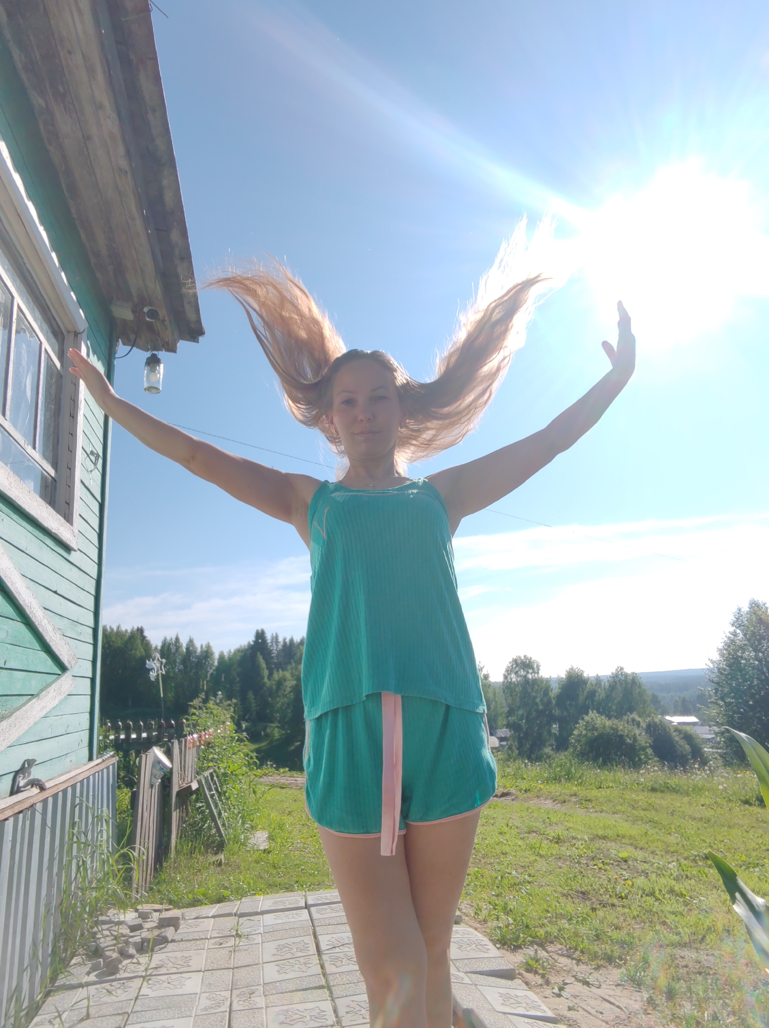 A young woman stands outside a wooden house in Чухлэм, Russia, with her arms outstretched and her long, light brown hair fanned out behind her, appearing as if caught by the wind. She is wearing a teal-colored top and matching shorts with pink trim. The sun is brightly shining, creating a lens flare and obscuring some details in the sky. To the left of the woman is the side of a teal-colored wooden building with a white-trimmed window and a weathered wooden roof. A light hangs from under the eaves. In the foreground, a tiled walkway leads away from the house. Beyond the walkway, there is a green lawn, a wooden fence, and a line of trees in the background under a clear blue sky.