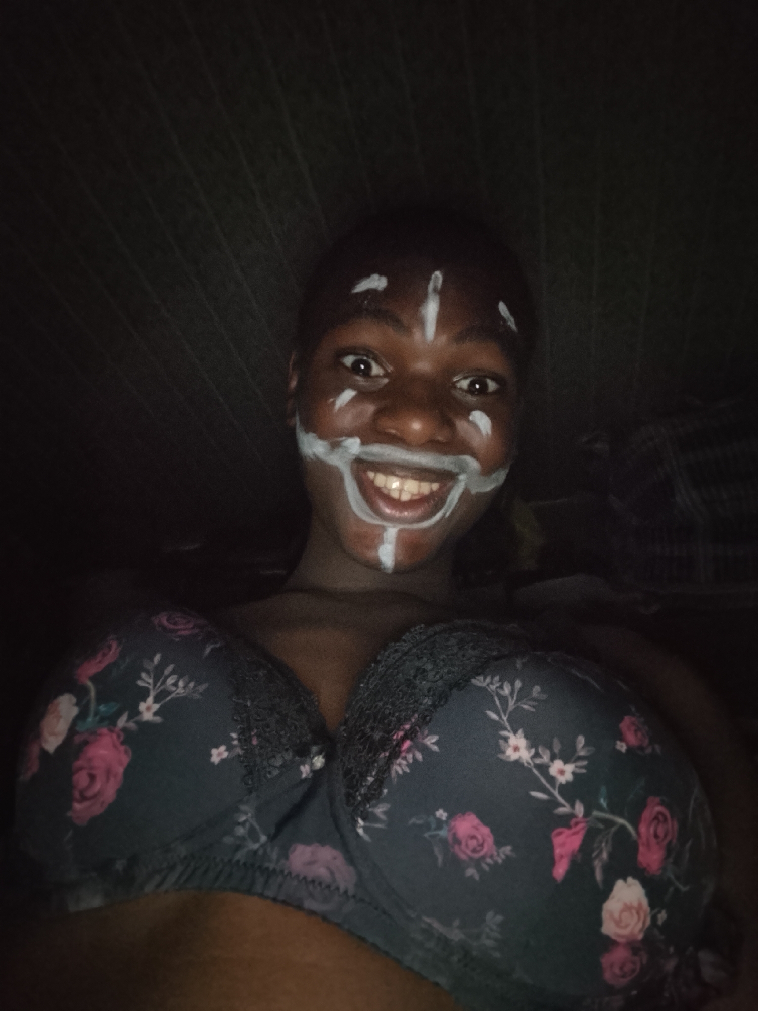 A dimly lit portrait shows a person with dark skin and short, dark hair, looking directly at the camera with wide, smiling eyes and an open-mouthed grin that reveals their teeth. Their face is adorned with white markings, including vertical lines on the forehead and under the chin, and horizontal lines across the cheeks, extending around the mouth in a playful manner. Small smudges or dots of white paint are also visible near the temples.
The person is wearing a dark bra featuring a prominent floral pattern of pink, red, and white roses with green leaves. The bra has lace detailing along the top edge of the cups. The upper torso and shoulders are visible from a low-angle perspective, suggesting a selfie.
The background is very dark and largely indistinct, with subtle textures possibly indicating fabric or a wall. The low lighting suggests an indoor setting, likely in the evening or at night. The light source appears to be directed from the front, illuminating the person's face and upper body. No text is visible in the image.