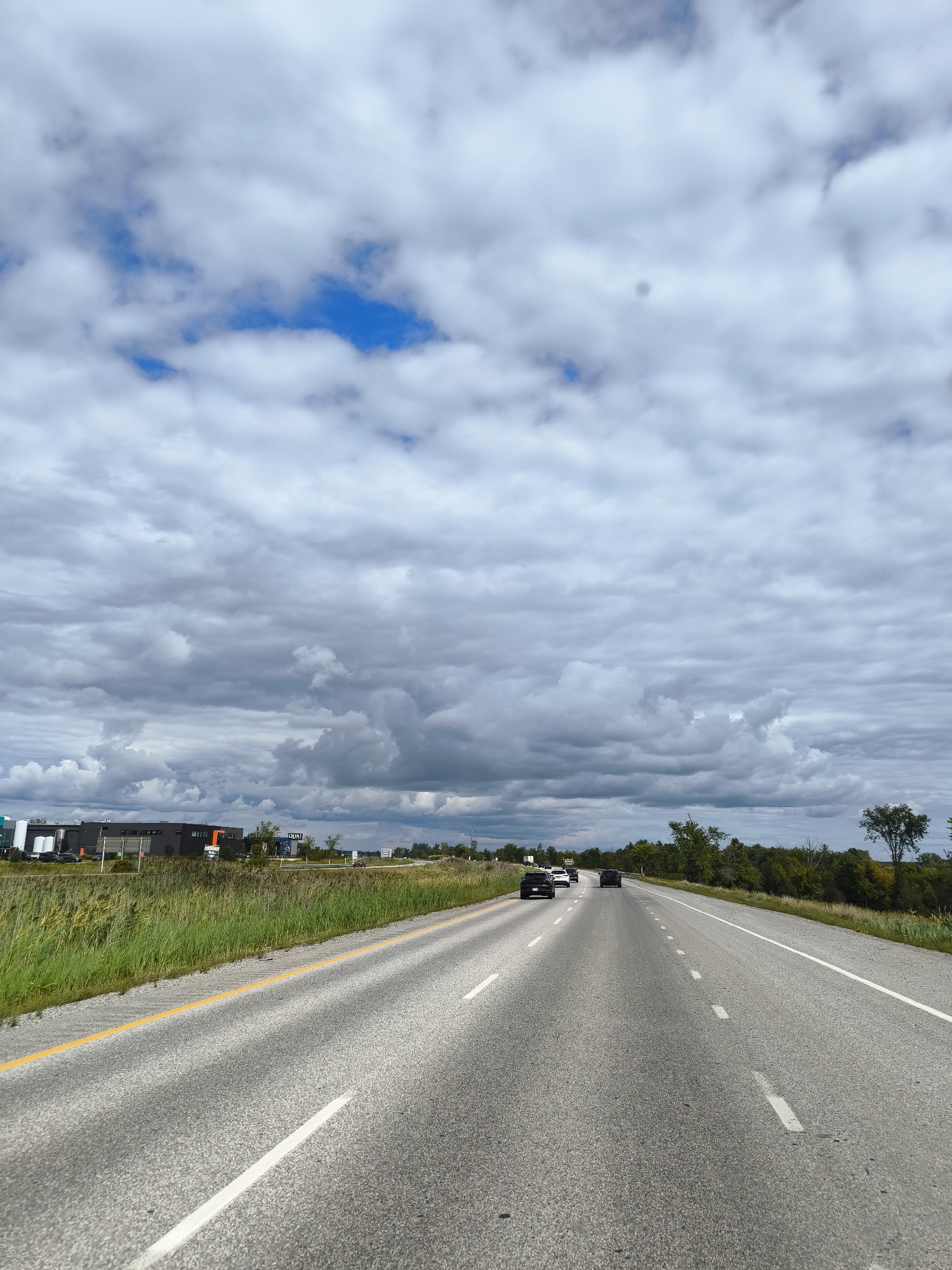 The image shows a highway with multiple lanes, a yellow line marking the left edge of the driving lane, and white lines indicating lane divisions. Several vehicles are visible on the road. To the left of the highway, there is a grassy verge, followed by a row of industrial-looking buildings and trees. The sky is filled with a mix of gray and white clouds, with patches of blue visible. The scene appears to be a daytime shot on a cloudy day, likely on a highway in an urban or suburban area, given the presence of industrial buildings. This image is of Highway 50, in Gatineau, Canada, heading east.