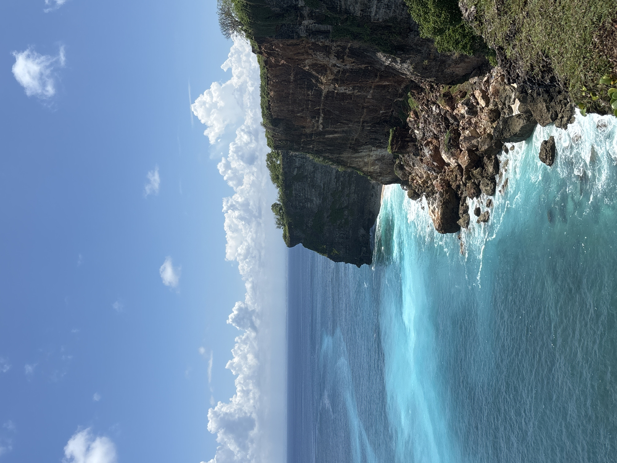 The image depicts a view from above of Uluwatu, Indonesia. A rugged cliff face, topped with green vegetation, plunges down to the turquoise ocean. Waves crash against the rocks at the base of the cliff. The sky is blue with scattered white clouds.