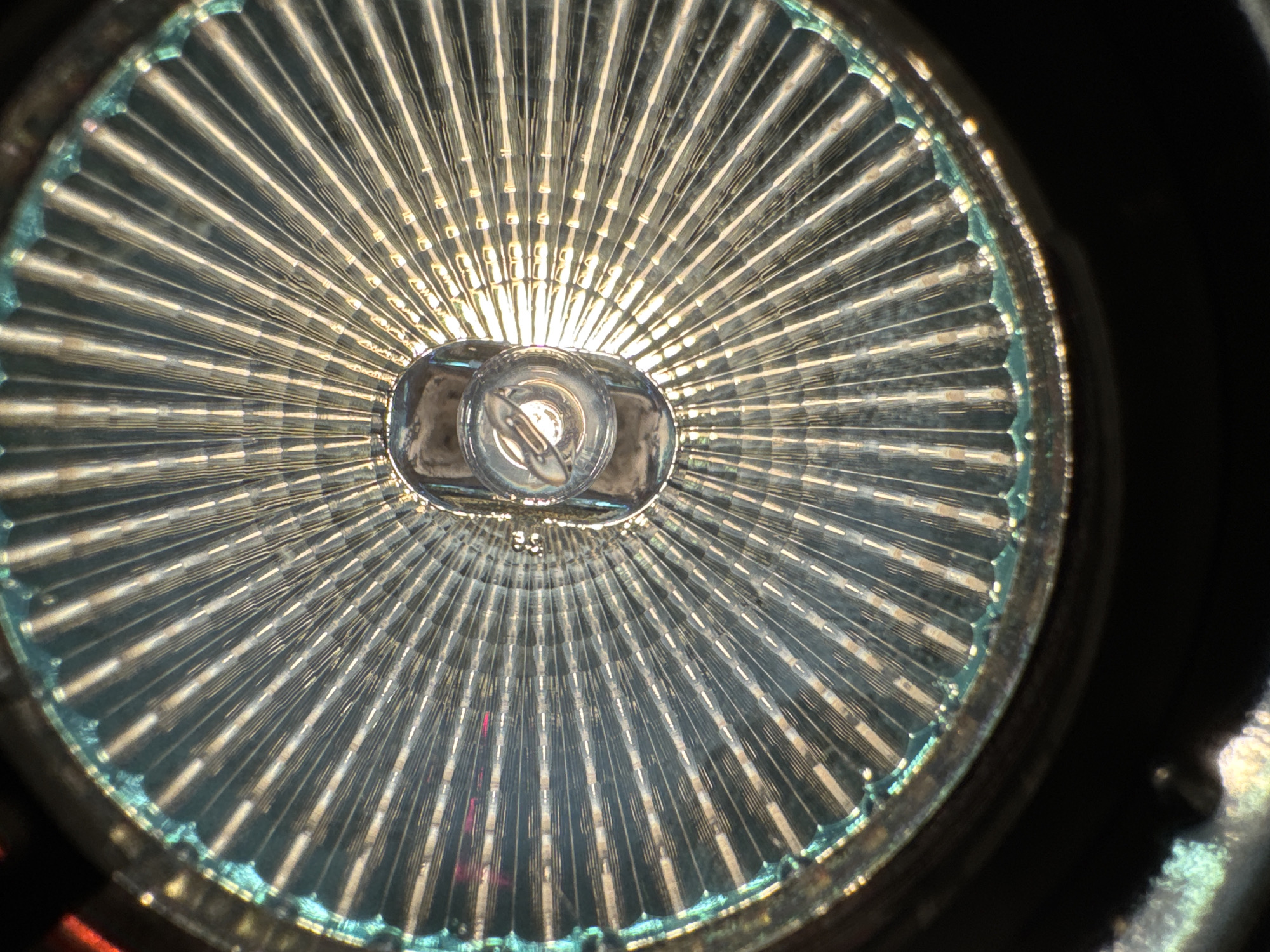 A close-up, overhead view of the reflector inside a flashlight. The reflector is made of glass with a patterned, ribbed surface that radiates from the center. At the center, a small, clear light bulb is visible, surrounded by a metallic housing. The outer edge of the reflector has a teal-colored, iridescent coating. The background is dark, suggesting the flashlight is either off or the surrounding area is unlit. The image does not provide any information about a specific location in the city of Unknown, Unknown.