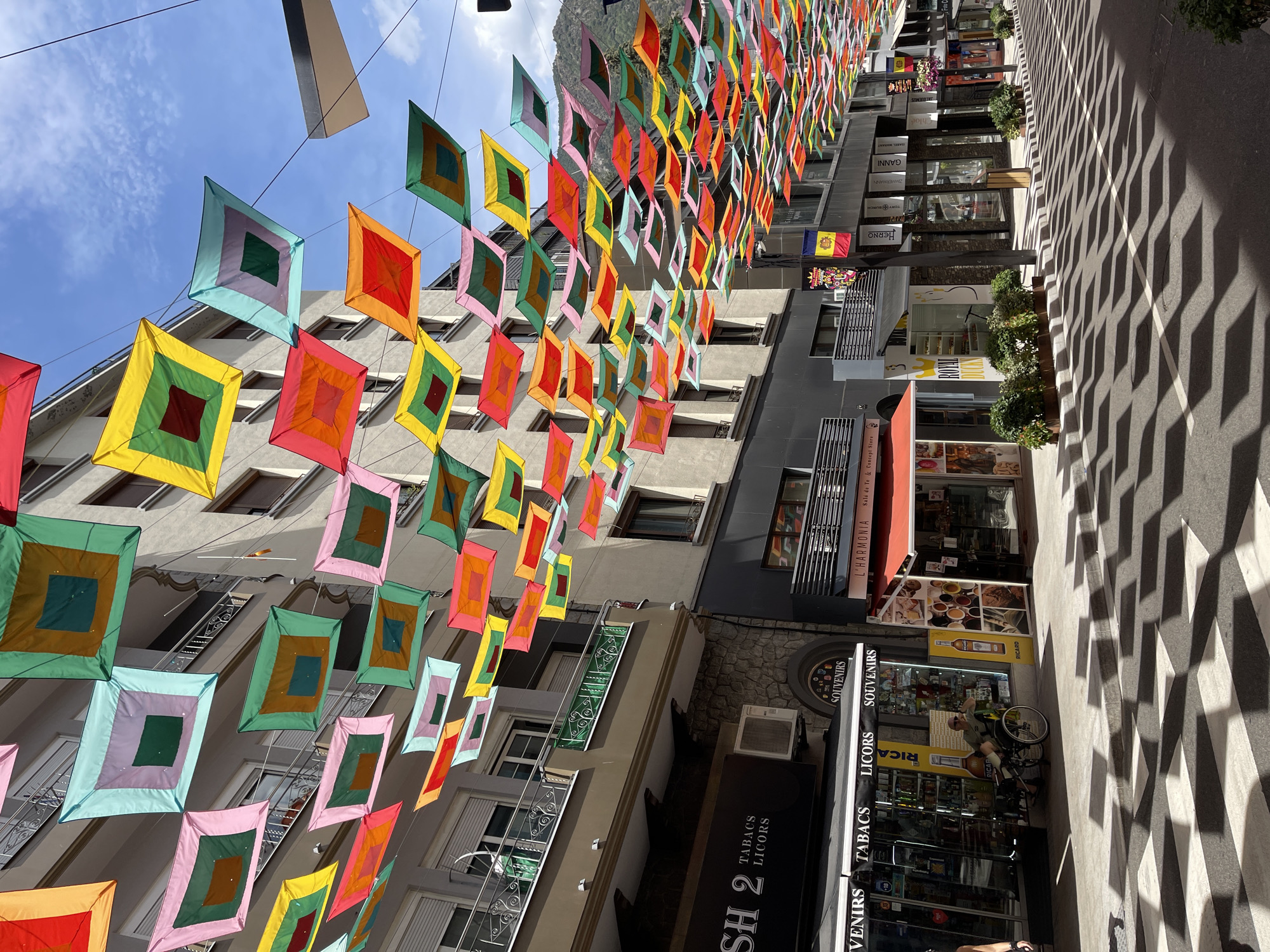 The image shows a street scene in a city, likely in Europe, with numerous colorful square fabric decorations hanging overhead. The decorations are arranged in rows and appear to be made of transparent or semi-transparent material in various colors including pink, green, yellow, orange, and blue, with darker square patterns in the center of some. The street is paved with a pattern of black and white stripes, likely a pedestrian crossing or part of the road design.
On the left side of the image, there are buildings with multiple stories featuring balconies with decorative railings. Shops are visible on the ground floor, with signs indicating "SH 2 TABACS LICORS SOUVENIRS" and "VENIRS TABACS LICORS SOUVENIRS". A person in a wheelchair is seated outside one of the shops.
On the right side of the street, there are more buildings with shops displaying their wares and store names like "L'HARMONIA". Flags, including what appears to be the flag of Andorra, are visible. The background shows buildings and hills or mountains, suggesting an urban environment situated in a mountainous region. The sky is blue with some clouds. The overall impression is a vibrant and festive street decorated for an event or season.