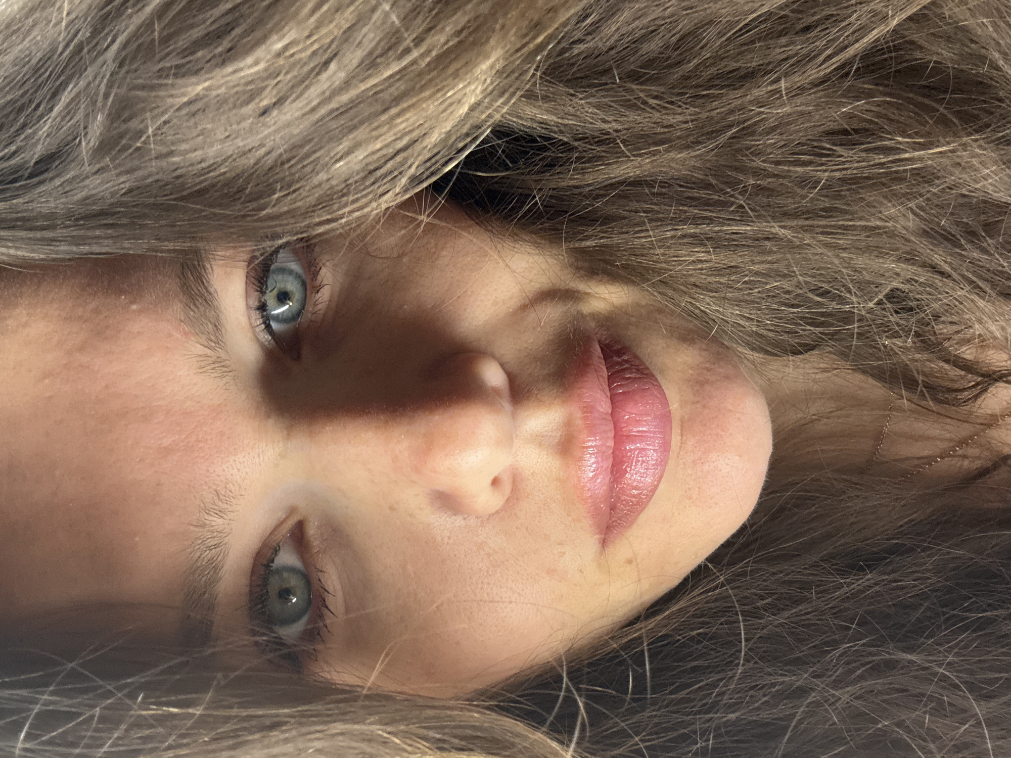 A close-up, portrait-style photograph depicts a person's face, predominantly focusing on the eyes, nose, and lips. The eyes are a light color, possibly blue or green, with visible eyelashes. The skin tone appears fair with freckles. The lips are slightly parted and have a glossy texture. The person is surrounded by strands of light brown or blonde hair, which frames the face and adds texture to the image. The orientation of the face is rotated, with the top of the head towards the left side of the frame. The background is obscured by the hair. There is no information to suggest the photograph was taken in Khimki, Russia.