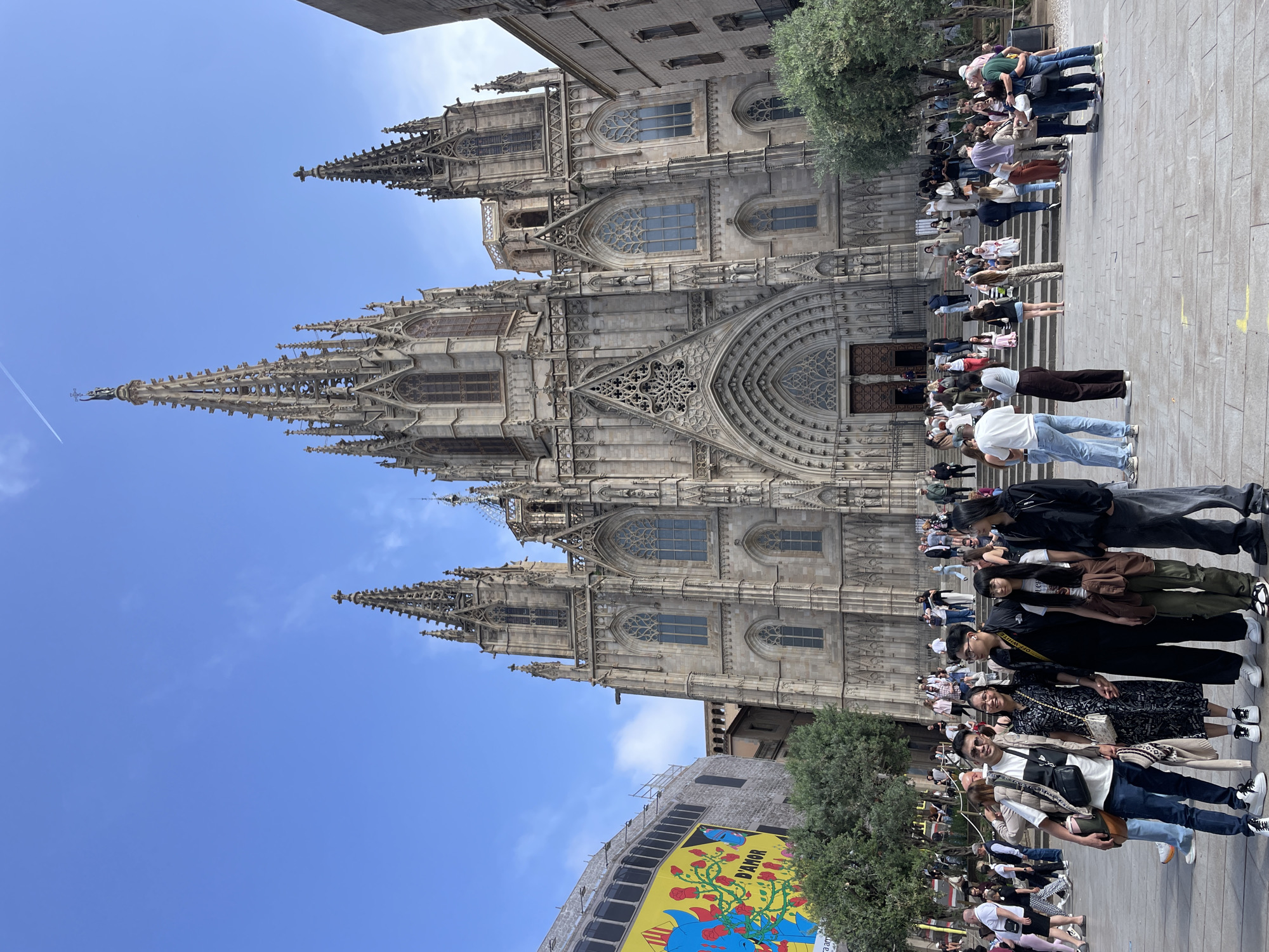 This daytime photograph captures the majestic Gothic architecture of a cathedral, identified as the Barcelona Cathedral, under a clear blue sky with a few wispy clouds and a faint airplane contrail. The cathedral's elaborate facade features multiple towering spires, intricate stone carvings, arched entrances, and large ornate windows.
In the foreground, a bustling plaza is filled with a large crowd of people, primarily appearing to be tourists or visitors. Many are seen strolling, observing the impressive structure, or queuing. Their casual attire suggests warm weather. In the immediate foreground, a group of individuals is posing and smiling for a photograph, contributing to a lively and cheerful atmosphere.
To the lower left of the frame, a modern building is partially visible, featuring a large, vibrant yellow billboard. The billboard displays artwork of red roses and a blue figure, with the prominent text "D'AMOR" clearly legible, along with some smaller, less distinct text that includes "33 aam". Green trees are interspersed within the scene, bordering the plaza. The image conveys a sunny and active scene at a historic landmark.
