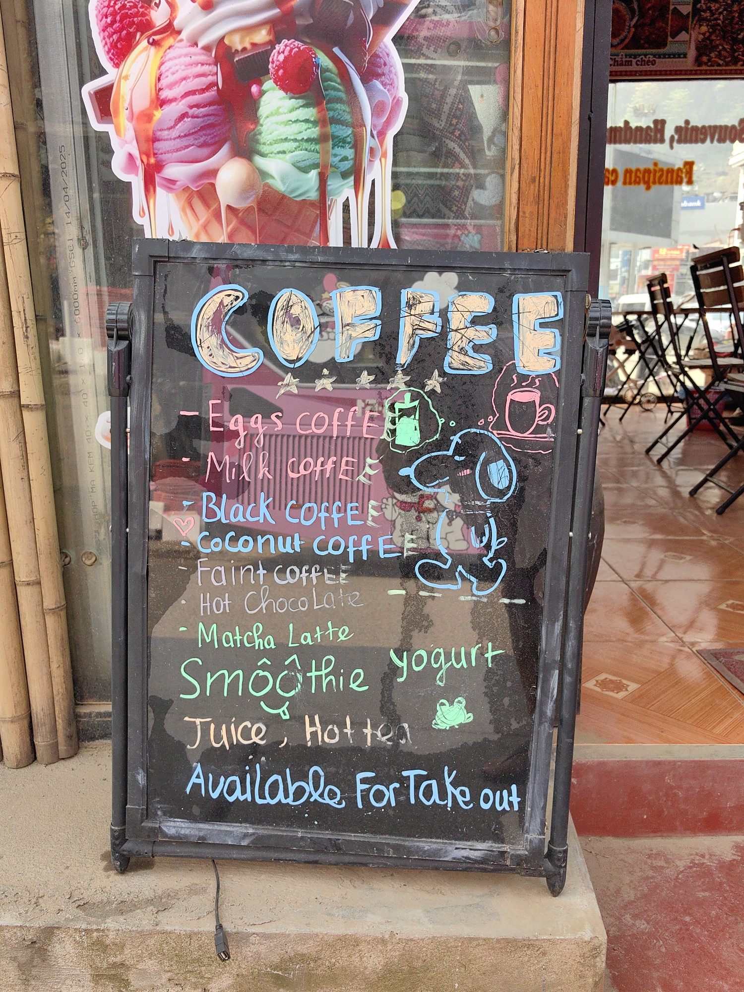 A black, double-sided, A-frame chalkboard menu board stands on a light brown concrete step outside a retail establishment in Sa Pa, Vietnam. The board displays hand-written menu items in various chalk colors, including "COFFEE" in large decorated letters, followed by "Eggs coffee," "Milk coffee," "Black coffee," "Coconut coffee," "Faint coffee," "Hot Chocolate," "Matcha Latte," "Smoothie," "Juice," "Hot tea," and "yogurt." The bottom of the sign states, "Available For Take out." Decorative chalk drawings include a light blue Snoopy-like dog character, a green frog, a pink hot beverage cup, a green iced beverage cup, and small yellow stars.
To the immediate left of the chalkboard is a glass storefront window featuring a large, colorful decal of an ice cream cone with three scoops (pink, green, and white soft-serve), topped with raspberries and chocolate drizzle. Reflections on this window include text such as "GSCL 14-04/2025" and "KEM 40 X 1.0". Several vertical bamboo poles are visible along the far left edge. To the right, a dark wooden door frame separates the left window from another storefront window. This right window reflects an outdoor seating area with dark, foldable tables and chairs. Reversed text, including "Souvenirs, Handmade," is visible in the reflection. The ground immediately beyond this doorway is tiled with brown and tan geometric patterns. A red-painted concrete step extends into the right foreground. A black cable, resembling a USB cable, dangles from beneath the menu board onto the concrete step. No individuals are present in the image.