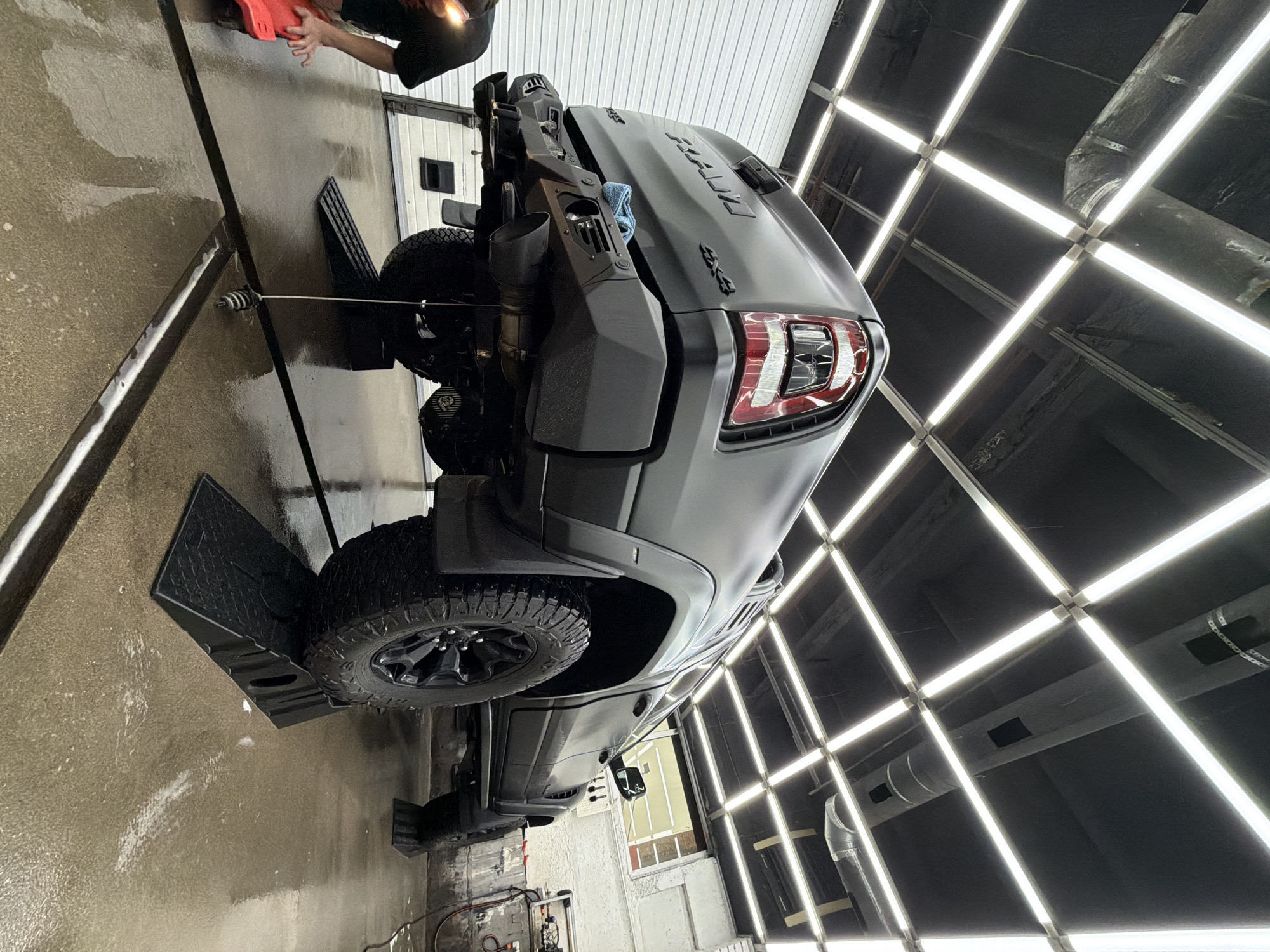 A low-angle, tilted medium shot shows a dark grey Ram TRX pickup truck being cleaned inside a car wash. The truck is elevated on ramps. A person, cropped at the shoulders, wearing a black t-shirt holds a red object in the upper left corner of the image. The car wash features a grid of bright lights on the ceiling. Hoses and cleaning equipment are visible on the floor in the lower part of the image. The scene is clean and well-lit. Based on visual features of the vehicle, environment, and general cleanliness, the image location cannot be confirmed as Krasnoyarsk, Russia.