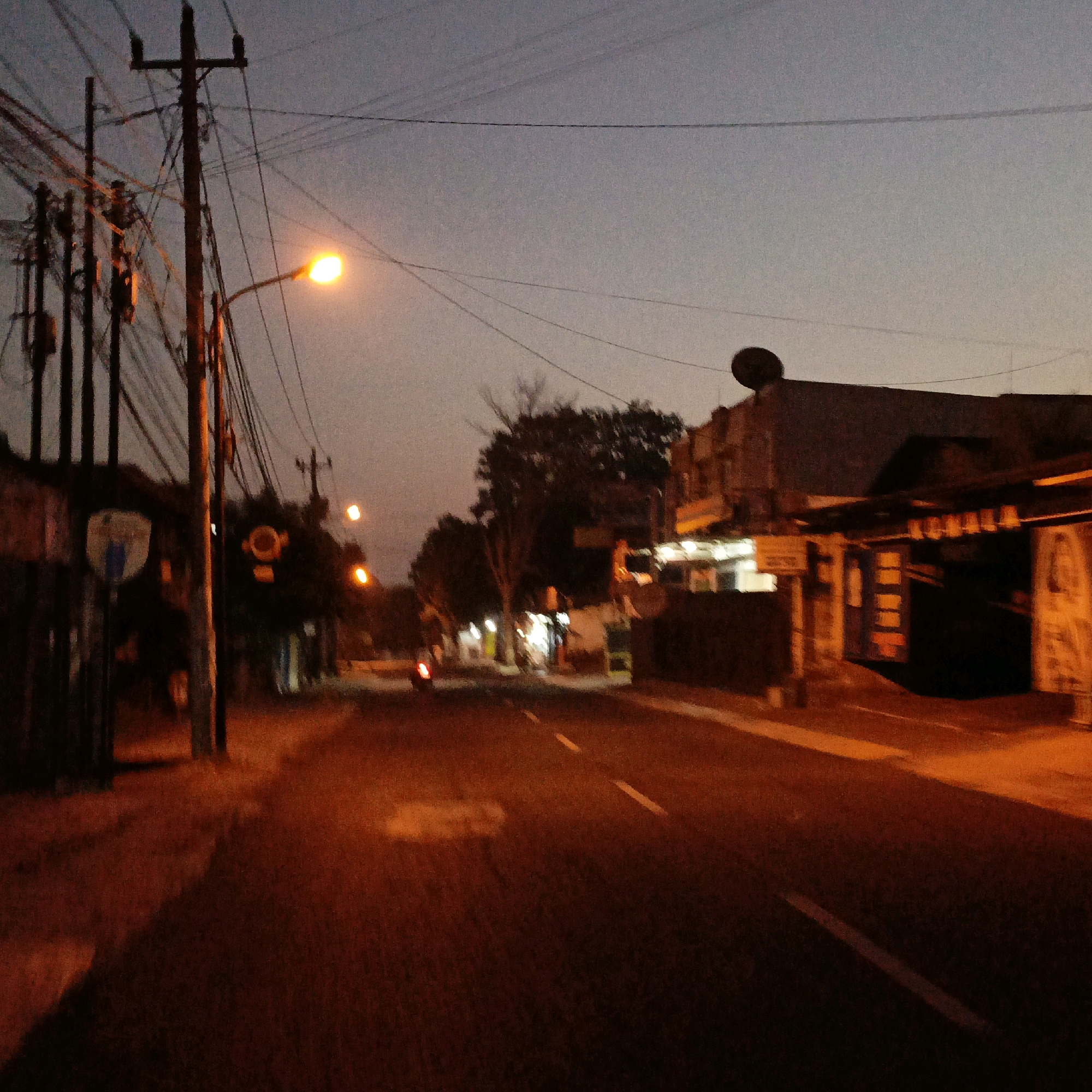 The image depicts a street scene at dusk in Salatiga, Indonesia. A street lamp on the left casts a warm orange glow on the asphalt road, which has white dashed lane markers. Utility poles with a complex network of wires crisscross the sky, which is a muted twilight blue. Buildings line the right side of the street, some with illuminated windows or signs. There's a faint silhouette of a motorcycle in the distance on the left. The overall impression is of a quiet, dimly lit urban environment transitioning from day to night.