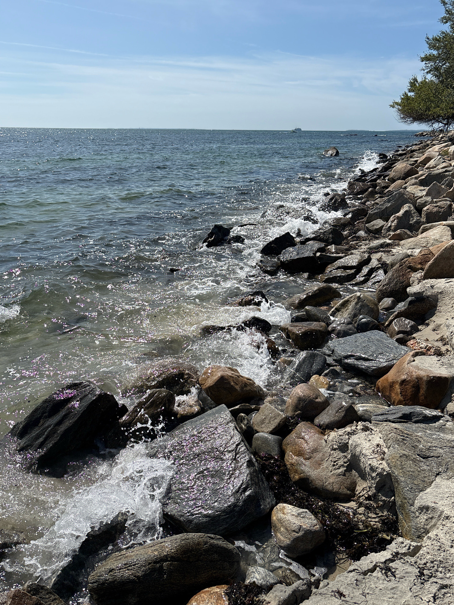 The image shows a rocky shoreline with waves crashing against the stones. The water is clear and reflects the sunlight, creating a shimmering effect. In the distance, a ship can be seen on the horizon. The sky is blue with a few scattered clouds. The shoreline is made up of a variety of rocks, some smooth and rounded, others rough and jagged. There is also some seaweed washed up on the shore. The overall scene is one of natural beauty and tranquility. This scene appears to be located on the coast of Waterford, Connecticut, USA.
