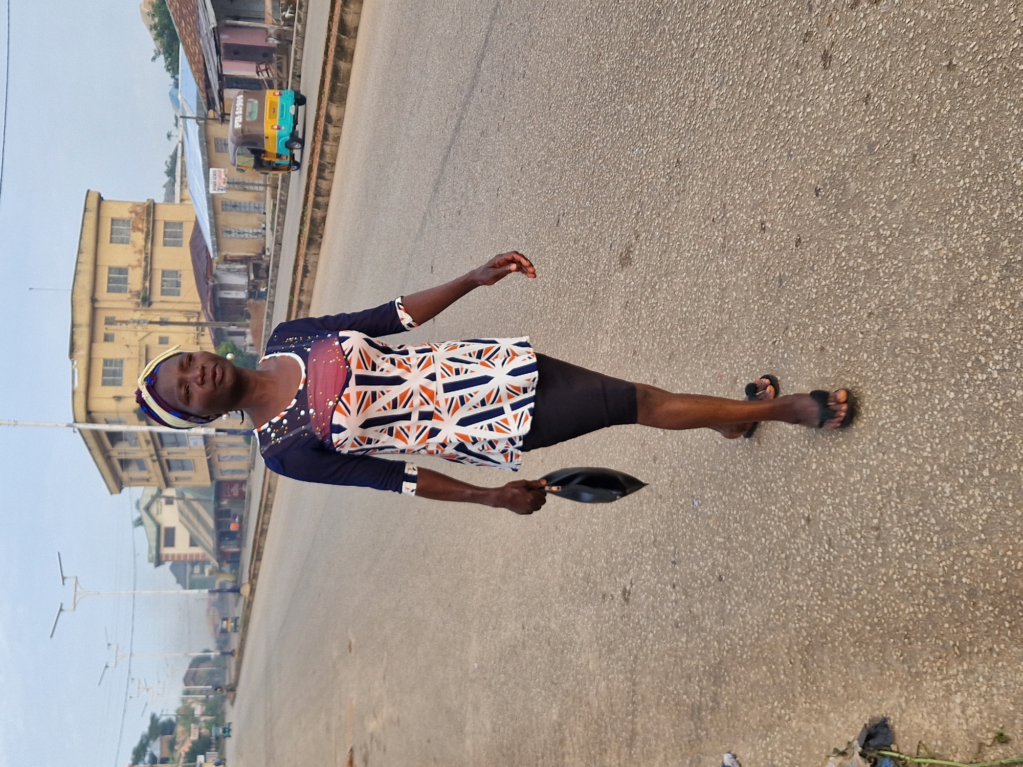 In Kabong, Nigeria, a woman walks down a street. She has dark skin and is wearing a colorful, patterned shirt and a headscarf. She is carrying a black handbag in her left hand. The street is paved and appears to be relatively empty, with a few buildings and a colorful auto-rickshaw visible in the background. The sky is overcast.