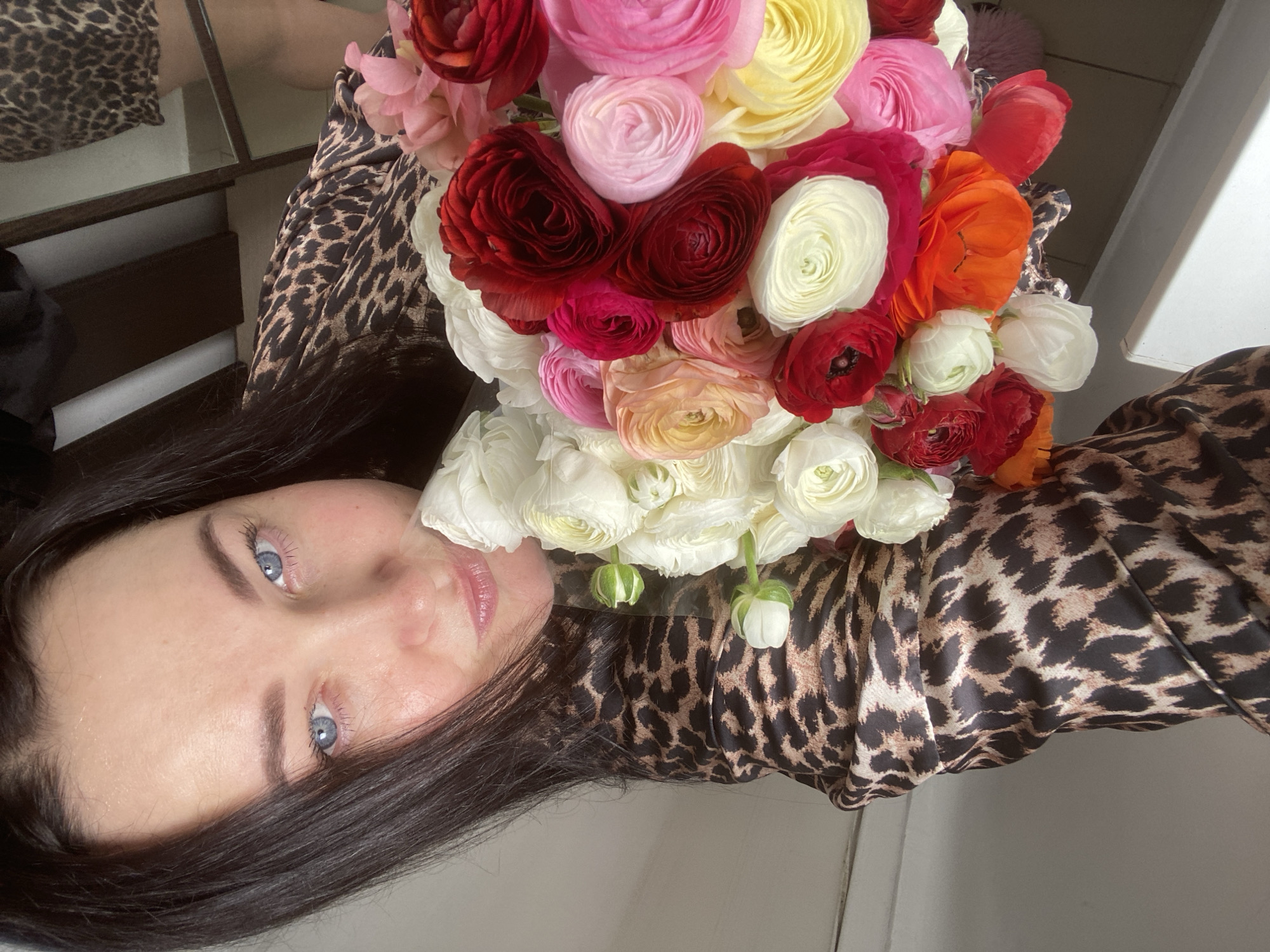 A woman with dark hair and light blue eyes poses while holding a bouquet of colorful ranunculus flowers. She is wearing a leopard-print top. The background includes a mirrored surface and a white wall. The location is not specified in the image.