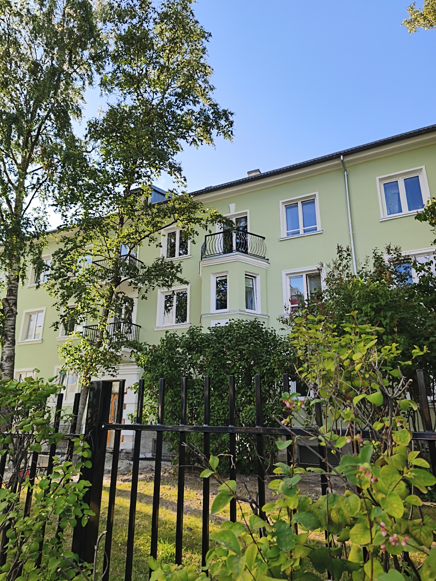In the city of Tallinn, Estonia, a three-story apartment building with a pale green facade is visible under a clear blue sky. The building features white-framed windows, some of which are reflective, showing the surrounding foliage. A balcony with a black metal railing juts out from the second floor. In front of the building, a black metal fence runs horizontally across the foreground, partially obscured by lush green bushes and trees. The foliage is dense, with some branches bearing small pink buds. The ground behind the fence appears to be a grassy area with some bare patches. The overall scene suggests a residential area bathed in natural light.