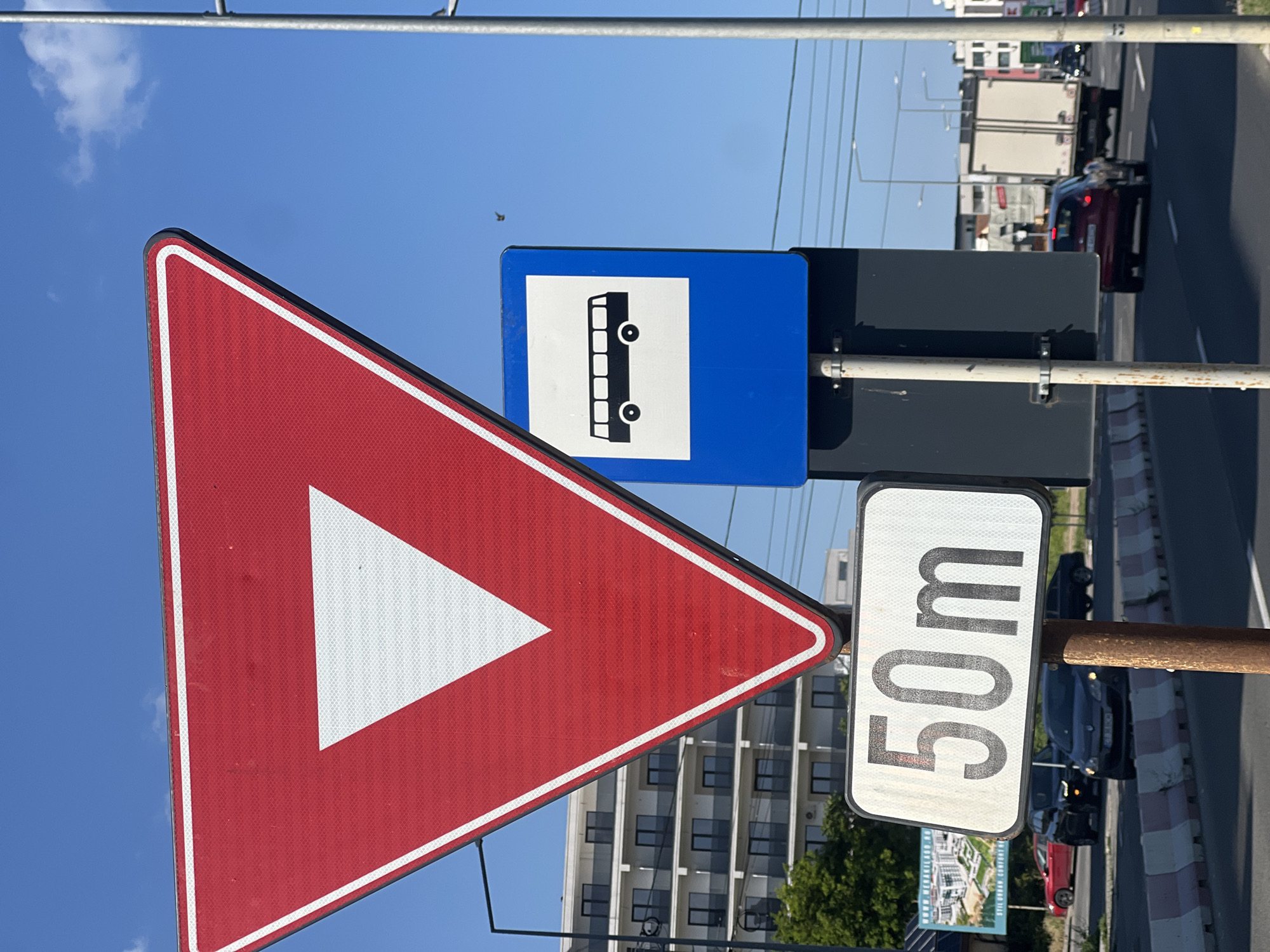 The image shows a street scene in Năvodari, Romania, on a clear, sunny day. In the foreground, three traffic signs are visible. The topmost sign is a red and white yield sign, shaped like an inverted triangle with a white triangle inside. To its right, a blue sign with a white silhouette of a bus indicates a bus stop. Below the yield sign, a rectangular white sign displays "50m" in black numbers and letters, likely indicating a distance. The signs are mounted on metal poles, one of which appears weathered and rusty.
In the background, a multi-story building with numerous windows and balconies is visible, suggesting a residential or commercial area. To the left, a billboard advertises "MEGA KILIAN RESORT" with an illustration of modern apartments and trees. The street is paved and marked with white lines. Several cars are on the road: a red sedan, a dark SUV, and a truck with a white cargo box. Traffic lights and street lamps are also present. The sky is a vibrant blue with a few wispy white clouds and a small bird in flight. The scene conveys a typical urban or suburban environment with active traffic.