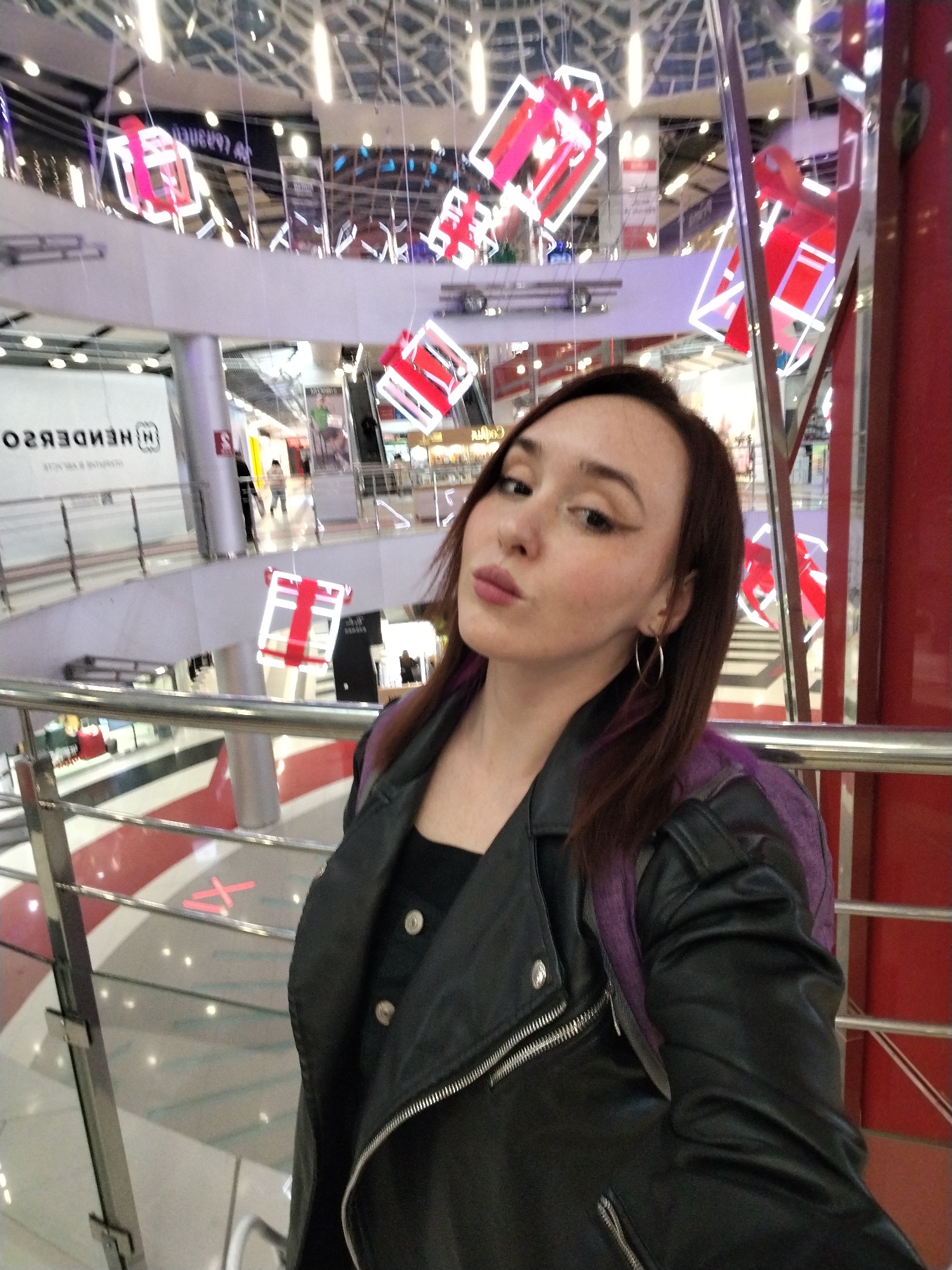The image is a selfie of a young woman taken indoors, likely within a multi-level shopping mall or large commercial complex in Izhevsk, Russia. The woman, who has medium-length brown hair with subtle purple undertones, is positioned in the foreground, looking directly at the camera with a slightly pouting expression. She is wearing a black leather biker jacket over a dark button-up top, and large silver hoop earrings are visible. A purple strap, possibly from a backpack, is visible on her right shoulder.
The background reveals the interior of a spacious building with multiple levels, connected by escalators or ramps. A white railing with silver posts separates the levels. Numerous decorative, illuminated abstract gift box structures, predominantly red and white, are suspended from the ceiling, suggesting a festive or holiday atmosphere. The ceiling features a grid pattern with bright lights.
Other individuals and faint shopfronts are visible on the lower levels. A prominent sign in the background displays mirrored text, "Н НЕНДЕВЯZО," which correctly reads "H HENDERSON" when un-mirrored, indicating a retail store. Red markings, including a large 'X', are visible on the floor of a lower level. The indoor lighting suggests a well-lit environment, possibly daytime or evening. The scene captures a casual moment during an outing in a modern commercial setting.