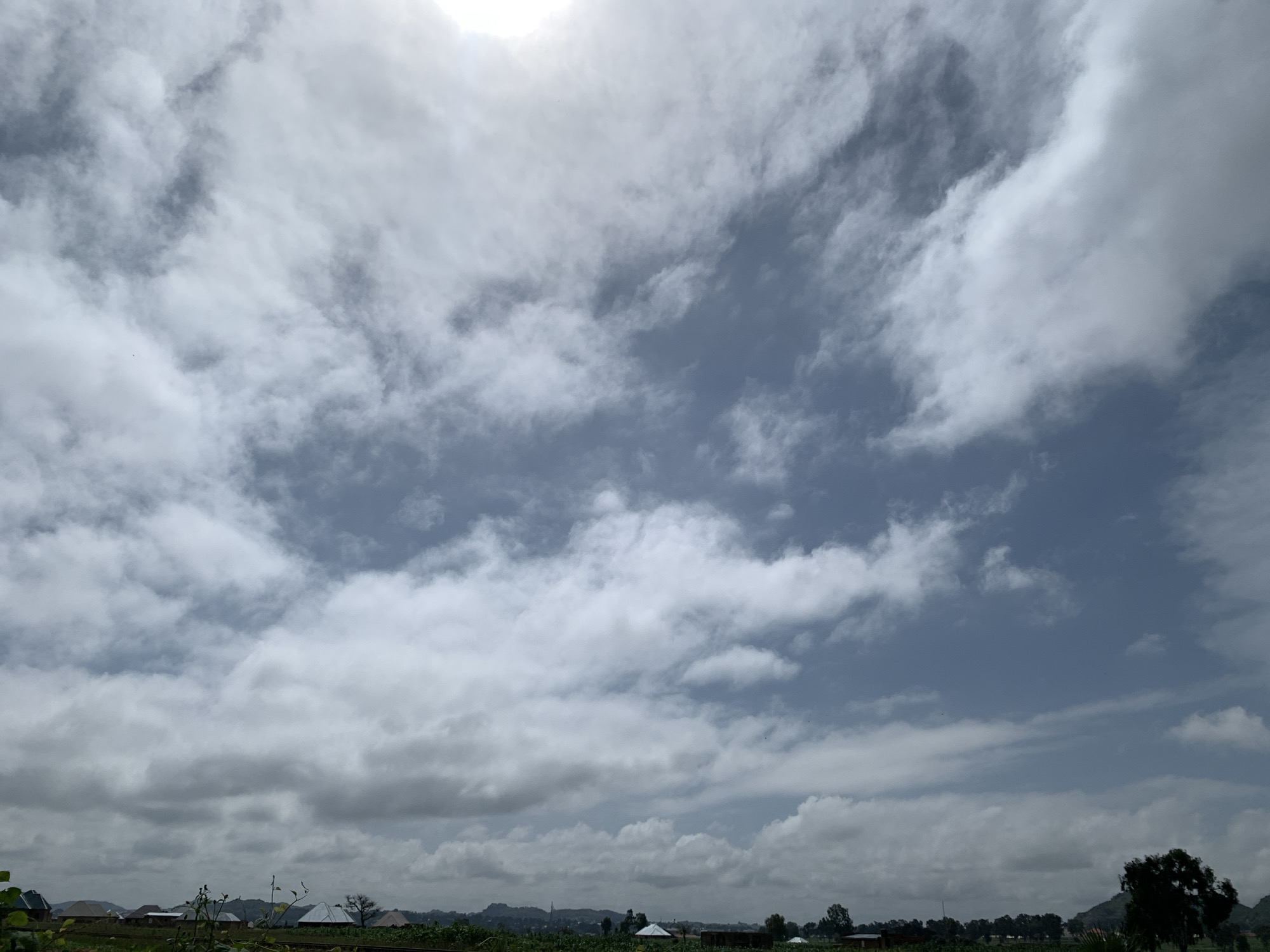 The image depicts a wide, overcast sky filled with dramatic, thick clouds that partially obscure the bright sunlight. A vast expanse of gray and white clouds stretches across the frame, with patches of a bright blue sky visible in some areas, especially towards the upper right. In the distance, a rural landscape is visible, characterized by scattered low-lying buildings with distinct roof shapes, some appearing to be residential structures with pitched roofs. Green vegetation, including trees and possibly fields, forms a lower band across the horizon. The scene suggests a quiet, possibly rural environment in the city of Kuru Karama, Nigeria, under a sky indicative of impending or recent rainfall. No people or active interactions are visible in the image.