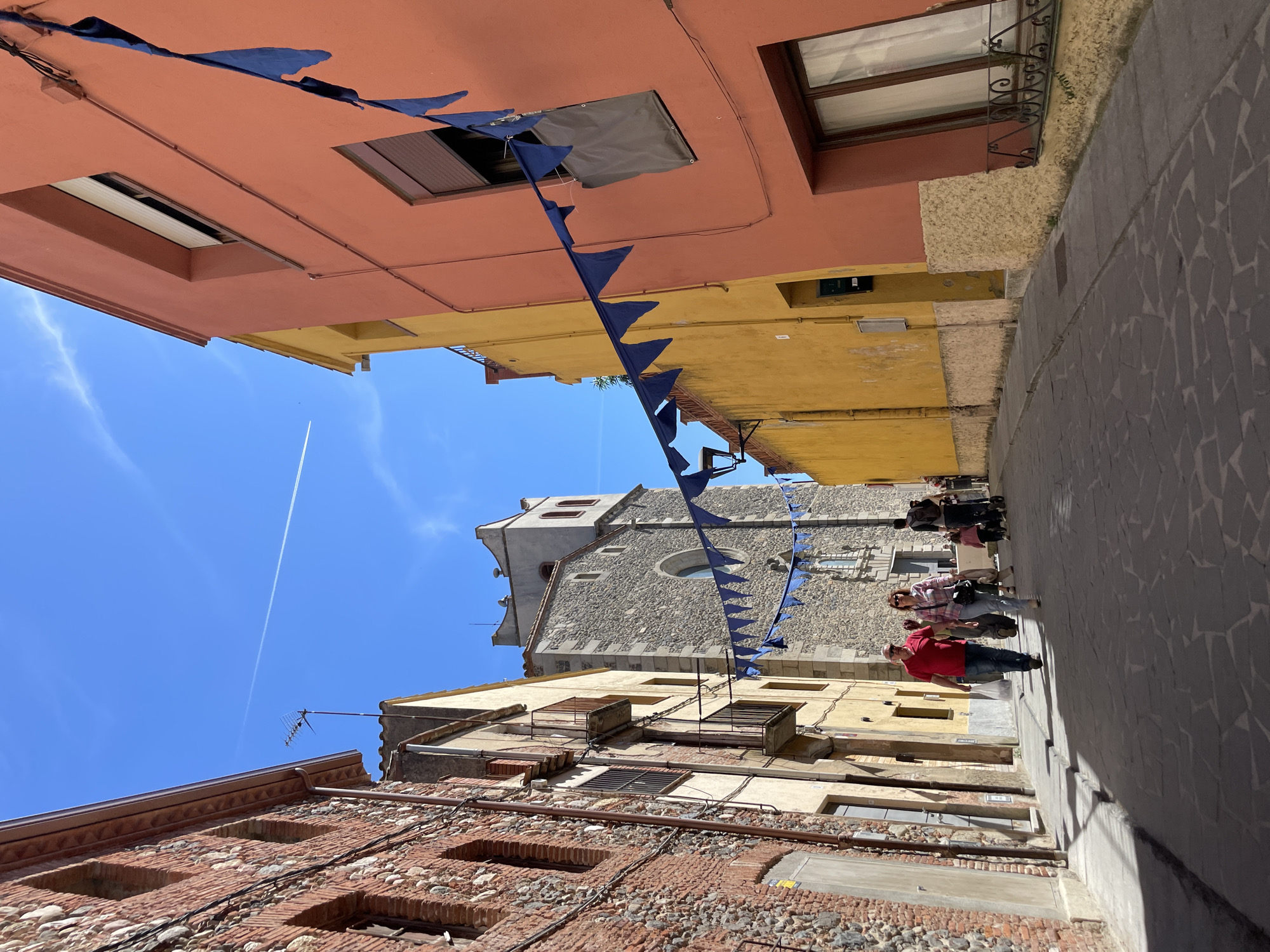 A low-angle shot in Hostalric, Spain depicts a narrow street scene with buildings of varying colors lining the sides. The buildings are constructed with brick and stone. Blue triangular banners are strung across the street overhead. Several people are walking along the street, including a man in a red shirt. A stone church tower is visible at the end of the street. The sky is blue, with a jet contrail visible. The street is paved with cobblestones.
