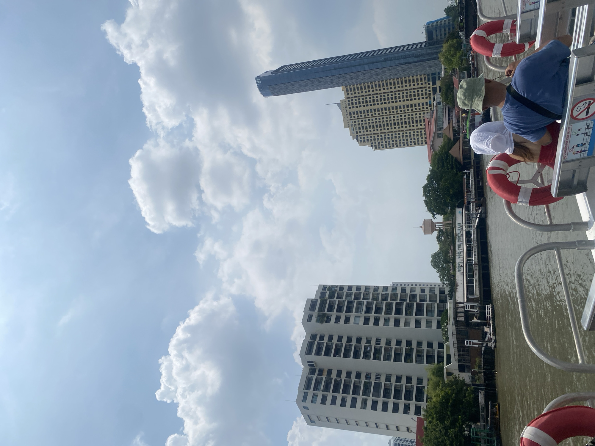 The image depicts a cityscape in Bangkok, Thailand, viewed from a boat on the Chao Phraya River. Two people, an older man wearing a green hat and blue shirt and a younger woman wearing a white hat and red shirt, are on the boat, leaning against the railing. Red and white life preservers are attached to the boat's railing. The river is visible, reflecting the buildings and sky. Prominent buildings include a tall, dark skyscraper (likely the Magnolias Waterfront Residences) and several other multi-story structures. Fluffy white clouds fill a large portion of the sky.