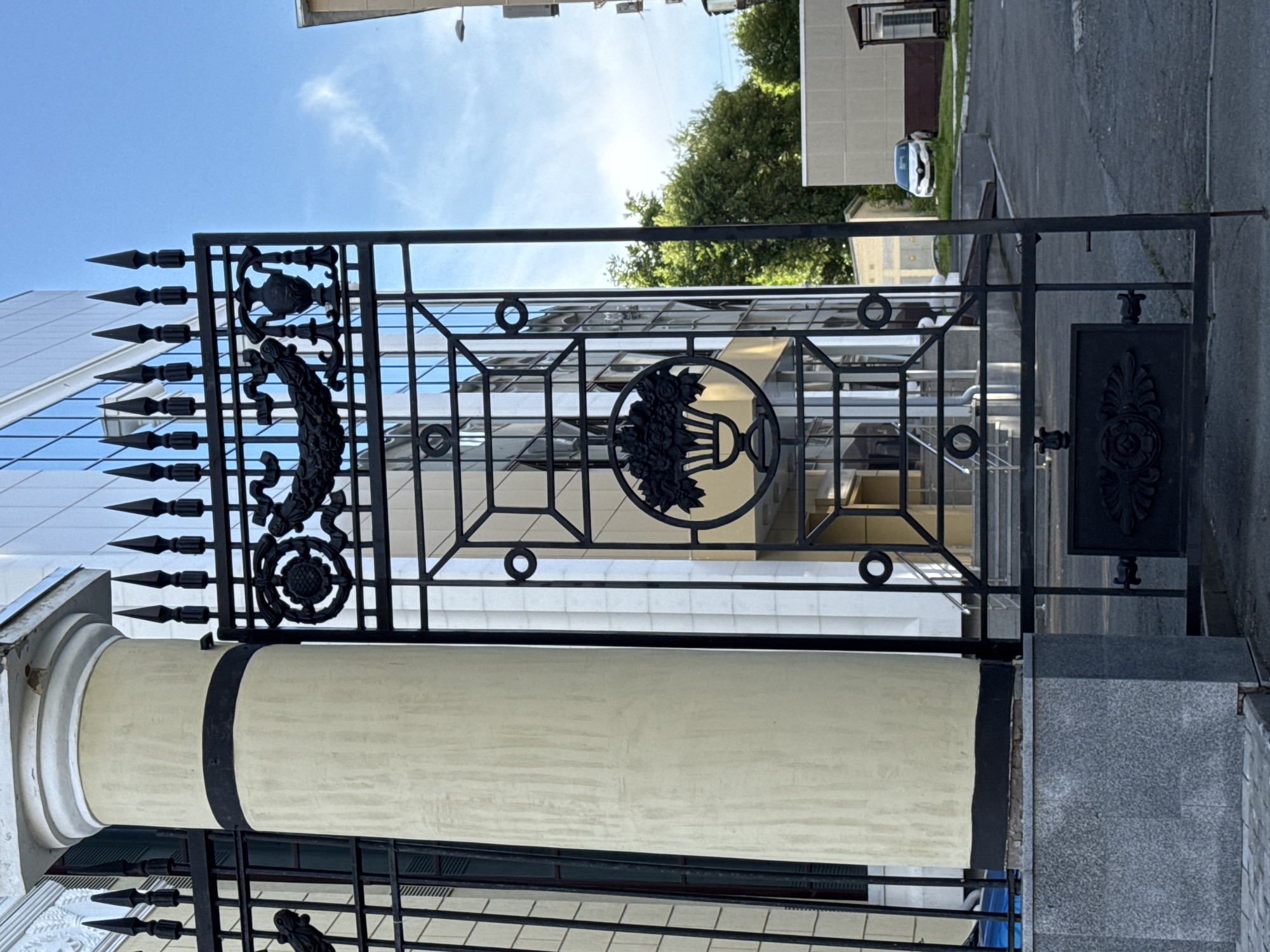 The image depicts a section of a black wrought-iron fence and gate in front of a modern building with a glass facade. The fence features ornate decorative elements, including a crest at the top left and a circular emblem with a floral design at the center. Spiked finials adorn the top of the fence. A large, cream-colored cylindrical column, possibly part of the building's entrance, is visible to the left of the gate.
In the background, the glass-paneled building reflects the bright blue sky and trees. Sunlight streams down, suggesting a clear and sunny day, likely midday. A parked car is visible on an asphalt surface to the right, beyond the fence, indicating an outdoor urban or semi-urban setting. The overall scene is composed of architectural elements and a bright, clear sky, conveying a sense of order and modernity. No people are visible in the frame. There is no discernible text in the image.