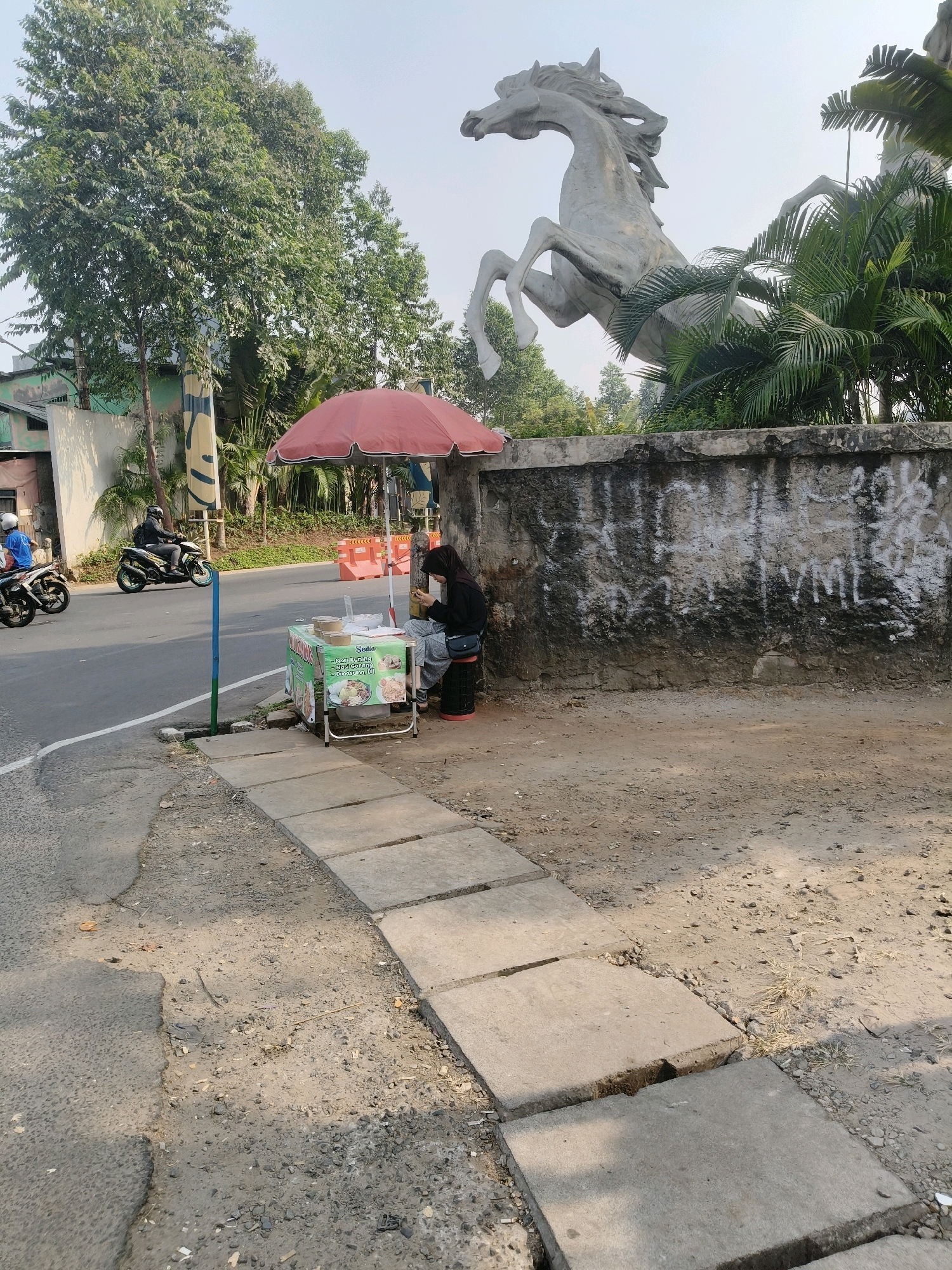 This image captures a street scene in South Tangerang, Indonesia, with a food stall as the focal point. A woman wearing a headscarf sits at the stall, which is covered by a red umbrella and displays various food items. Behind the stall, a concrete wall with graffiti is visible, and beyond that, a large sculpture of a horse stands against a backdrop of trees and palm fronds. The street itself is paved, with several motorbikes passing by, indicating active traffic. The overall environment suggests a sunny day, with distinct shadows cast on the ground.
