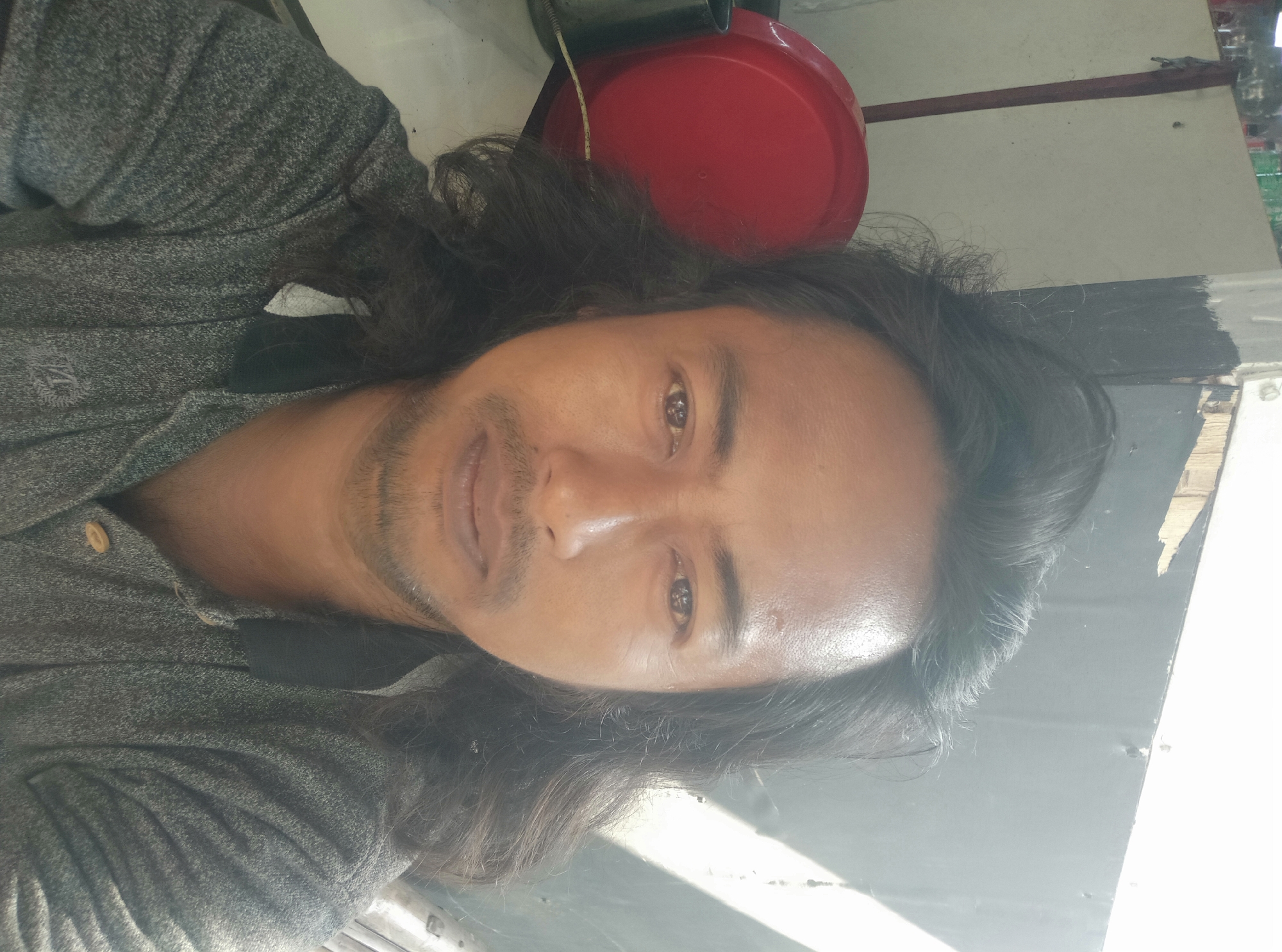 A medium shot selfie shows a man with long, dark, wavy hair and a light beard. He is wearing a grey polo shirt with a dark collar. The background appears to be an outdoor setting with a wall and sunlight casting shadows. A red bowl is visible to the left of the man. The image was taken in Sumurber, Indonesia.