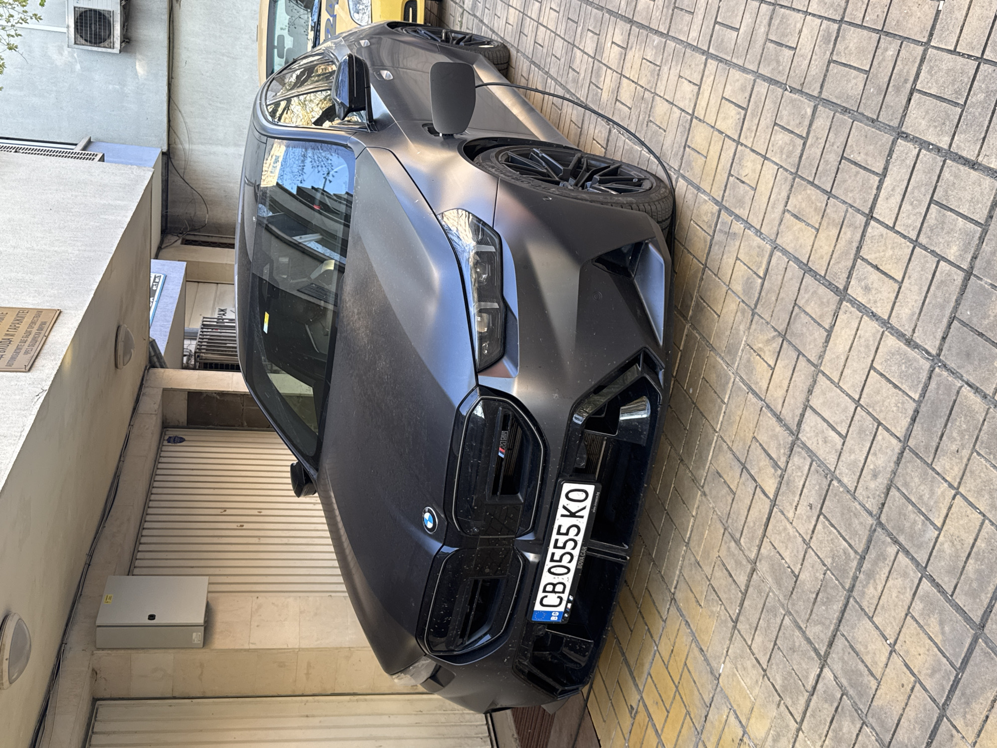 The image shows the front of a matte black BMW M5 parked on a brick-paved surface. The car has a Bulgarian license plate "CB 0555 KO" and a BMW logo is visible on the hood. The car is positioned close to a building with a garage door. A sign is seen on the building, which includes the text "ГАРАЖИТЕ" (GARAGES) in Bulgarian. Based on the license plate and signage, the location is likely in Burgas, Bulgaria.