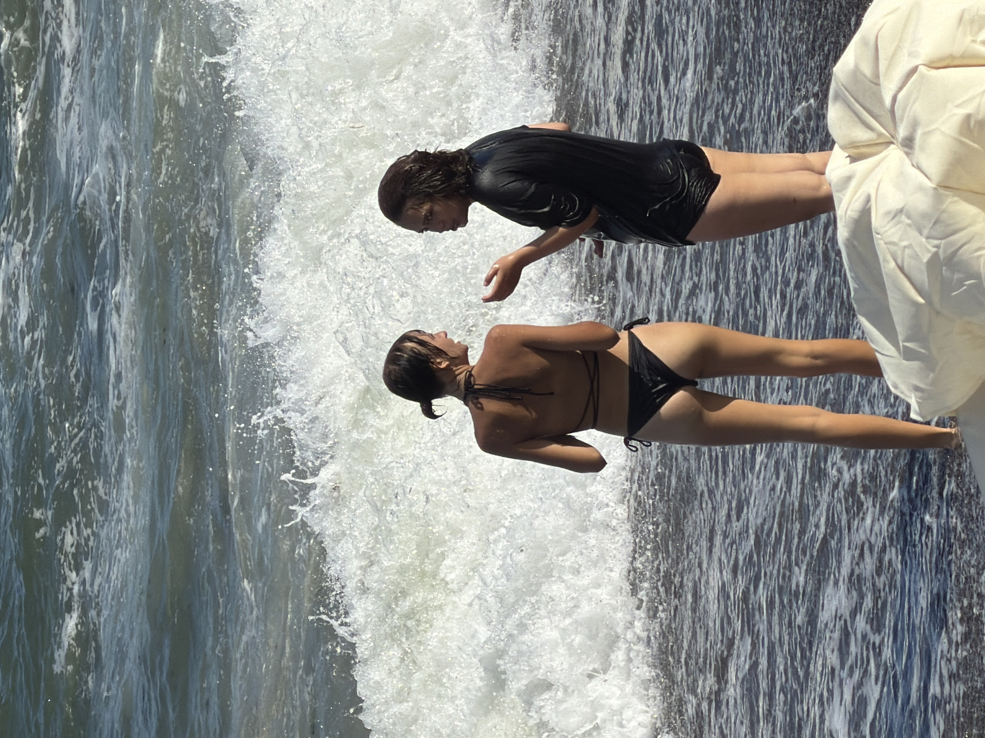 The image shows two young women on the shore of the sea in Alushta, Ukraine. The woman on the left is wearing a black bikini, and the woman on the right is wearing a black wet t-shirt. They are standing in shallow water with waves crashing behind them. The water is dark and the waves are white and foamy. There is a white object, possibly a beach towel or cushion, in the foreground. The sky is not visible in the frame.