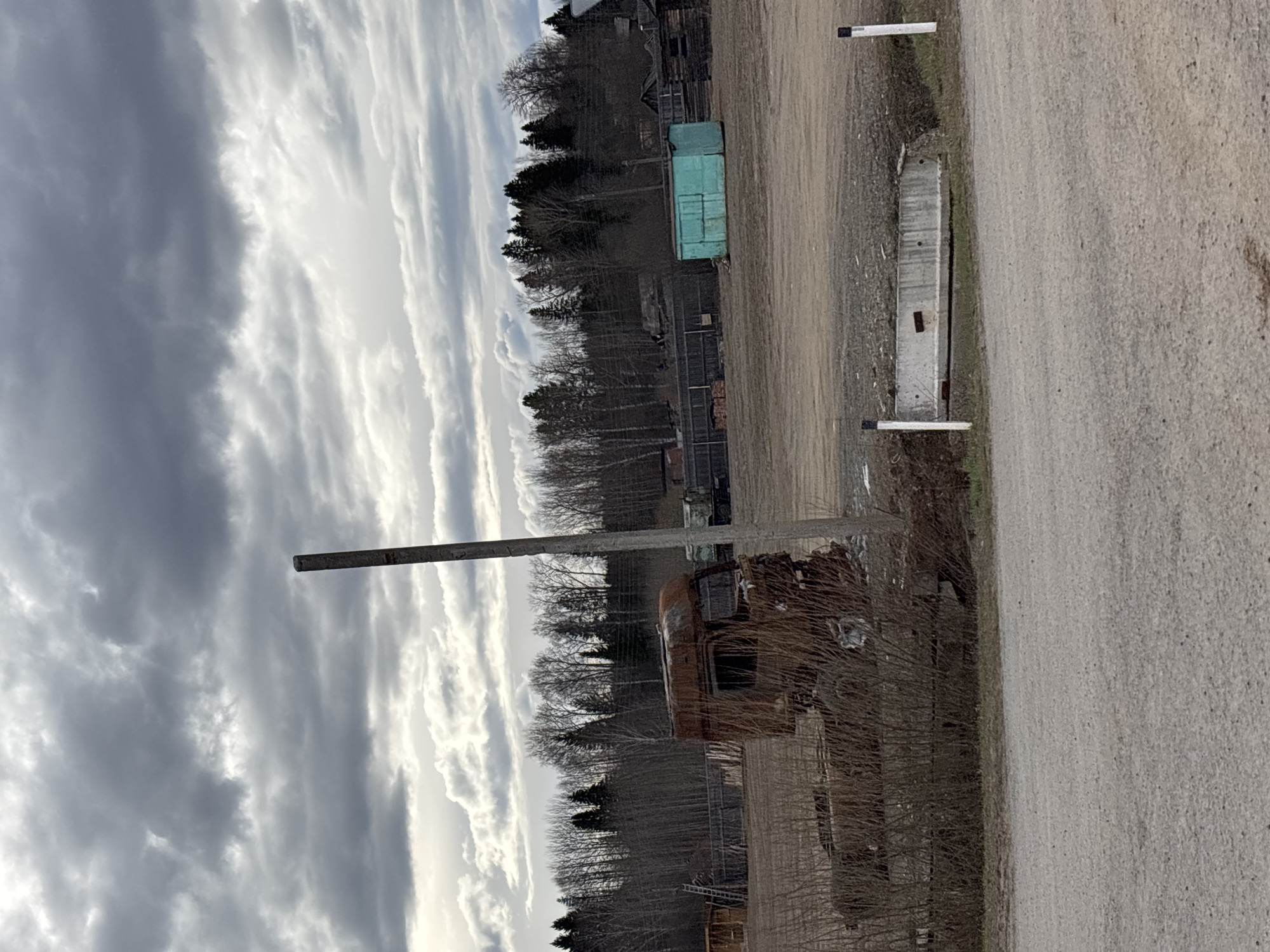 The image depicts a rural scene in Zachernaya, Russia, under a sky filled with textured, striated clouds. A dirt road runs vertically along the right side of the image. Adjacent to the road, a concrete structure is visible, marked at either end by what appears to be thin, white posts. The roadside transitions into a field where a rusted vehicle stands amidst dry vegetation. A tall, gray pole rises from the field towards the sky. Beyond the field, a line of bare trees defines the horizon, behind which is a dark, forested area. A green container and other dilapidated structures are vaguely discernible behind the trees. The overall scene presents an impression of dereliction and disuse.