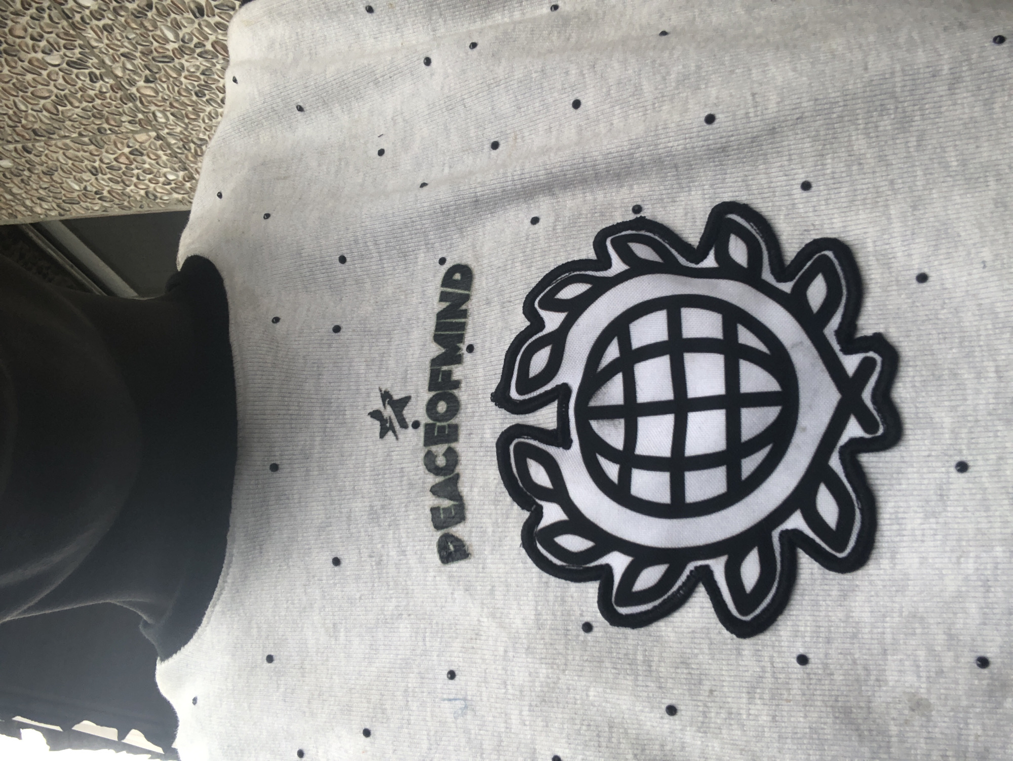 The image shows a close-up of a person wearing a light gray t-shirt with a black neckline. The t-shirt is decorated with small black dots scattered across the fabric. Centered on the chest is a black and white emblem. Above the emblem, the words "PEACEOFMIND" are embroidered in black lettering, with a small star graphic to the left of the text. The emblem itself features a stylized globe surrounded by a floral or leaf-like pattern. The background appears to be a textured wall, possibly made of small stones or pebbles. The lighting suggests an outdoor setting. The person's face and neck are visible, with the face obscured by shadow. The overall impression is a casual portrait focusing on the graphic design of the t-shirt. The location is identified as Ado, Nigeria.