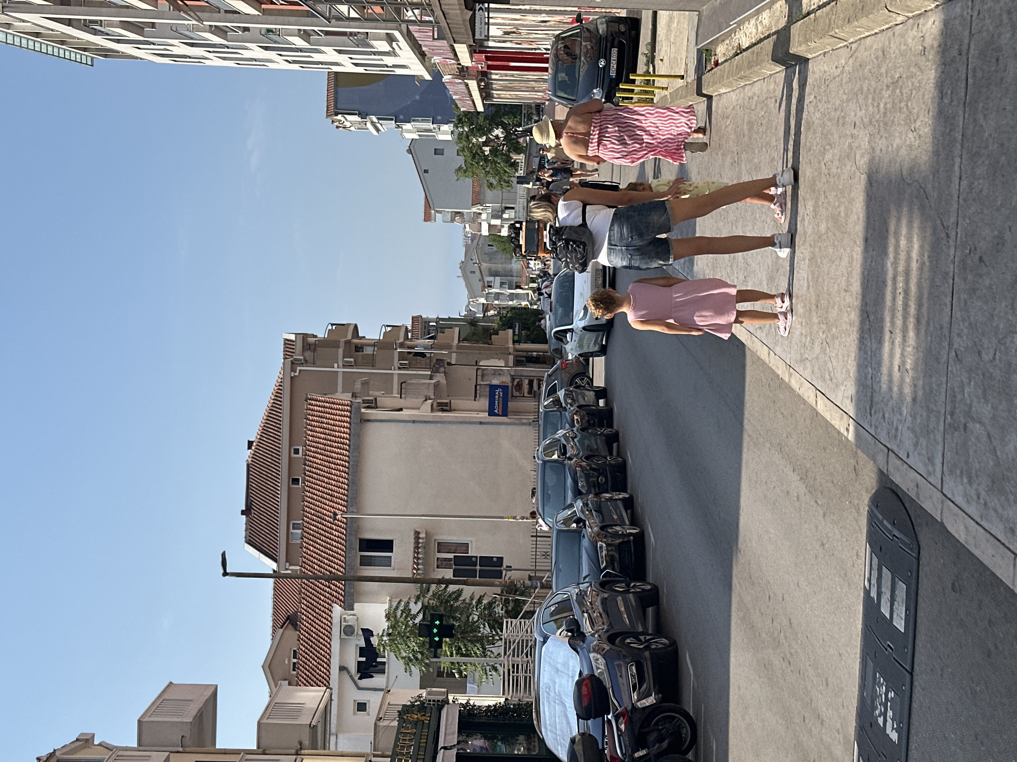 The image depicts a street scene in Budva, Montenegro on a sunny day. In the foreground, two young girls and an adult woman are walking on a sidewalk. The girl on the left wears a pink dress, and the girl on the right wears a yellow dress. The adult woman is wearing denim shorts and a white t-shirt, carrying a backpack. Another woman in a pink and white striped dress and a straw hat is walking ahead of them. The sidewalk is paved with concrete and has a curb.
The street is lined with parked cars, including several dark-colored SUVs and sedans. A motorcycle is also visible parked on the side. Across the street, buildings with terracotta tile roofs and balconies are visible, typical of Mediterranean architecture. Shops and businesses with awnings are situated along the street. A traffic light with green signals is illuminated. The sky is clear and blue. The overall scene suggests a bustling urban environment during daylight hours.