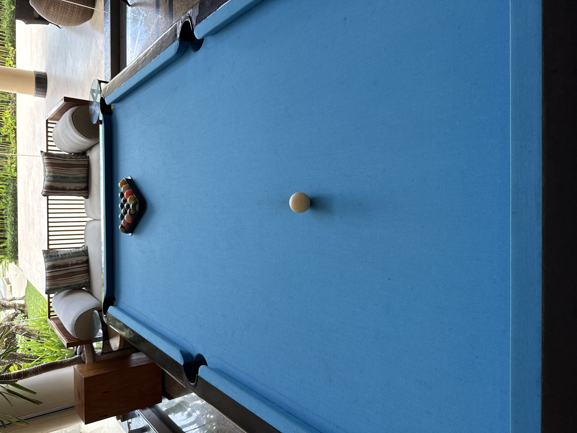 This image captures a pool table with a light blue felt surface. The billiard balls are arranged in a rack near the left edge, while the cue ball sits in the center of the table. The table's trim is dark, and its pockets are visible at various points along the edges. Adjacent to the table, a cushioned seating area with pillows is present. Beyond that is an outdoor area with landscaping and a glimpse of architecture. Based on the surroundings and the context provided, the location is Nusa Dua, Indonesia.