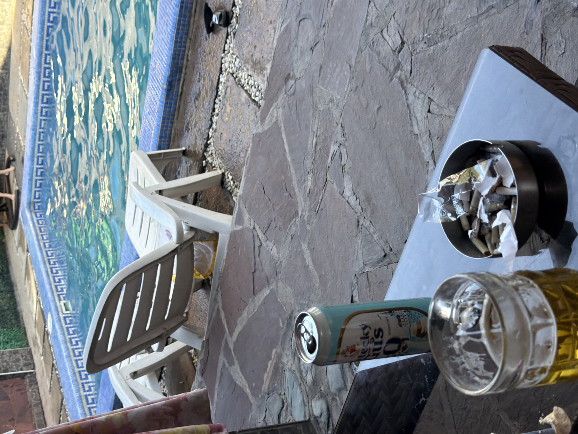 The image shows a close-up of a glass of beer and an open can of "Becky Gus" beer on a table next to an ashtray filled with cigarette butts. In the background, a swimming pool with blue tiles and clear water is visible, along with white lounge chairs. The scene appears to be an outdoor patio area, likely in a private residence or a hotel, suggesting a leisure setting. The presence of the beer and ashtray indicates that someone was recently relaxing or socializing. The overall ambiance is one of casual relaxation. The location is the city of Shymkent, Kazakhstan.