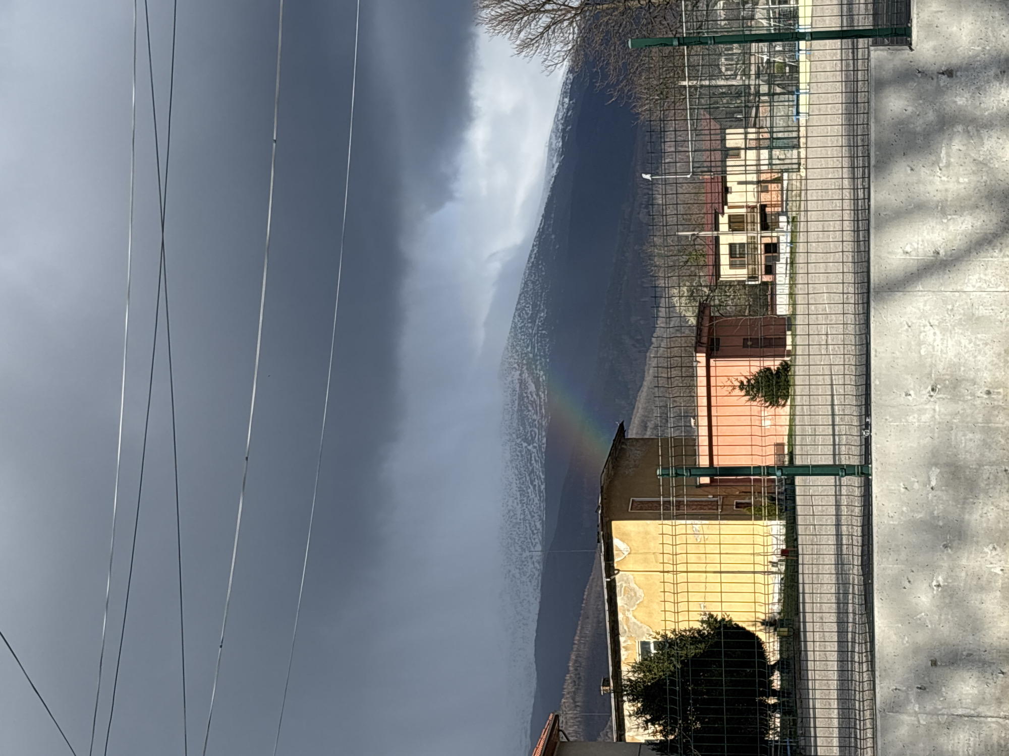 The image shows a landscape in Zlatitsa, Bulgaria, viewed from behind a wire fence and a concrete wall. Power lines are visible in the sky. The sky is overcast with dark clouds on the left. A rainbow is visible over a mountain in the distance. Several buildings are visible behind the fence. The road is on the right.